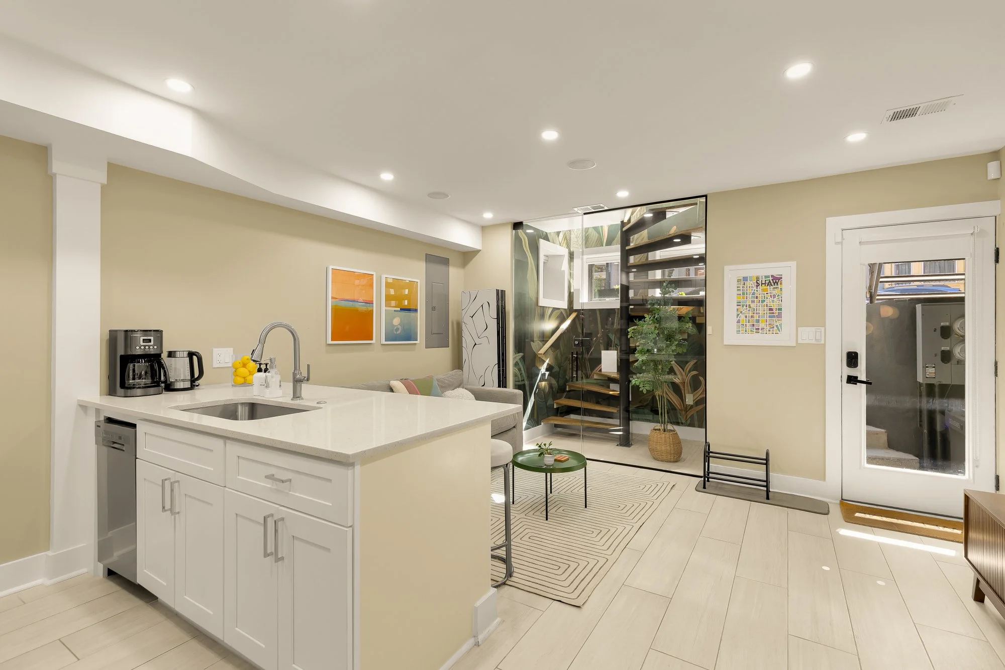 Modern kitchen and living area with a white kitchen island, artwork on the wall, a staircase with black and wooden steps, a door with a window, and decorative plants.