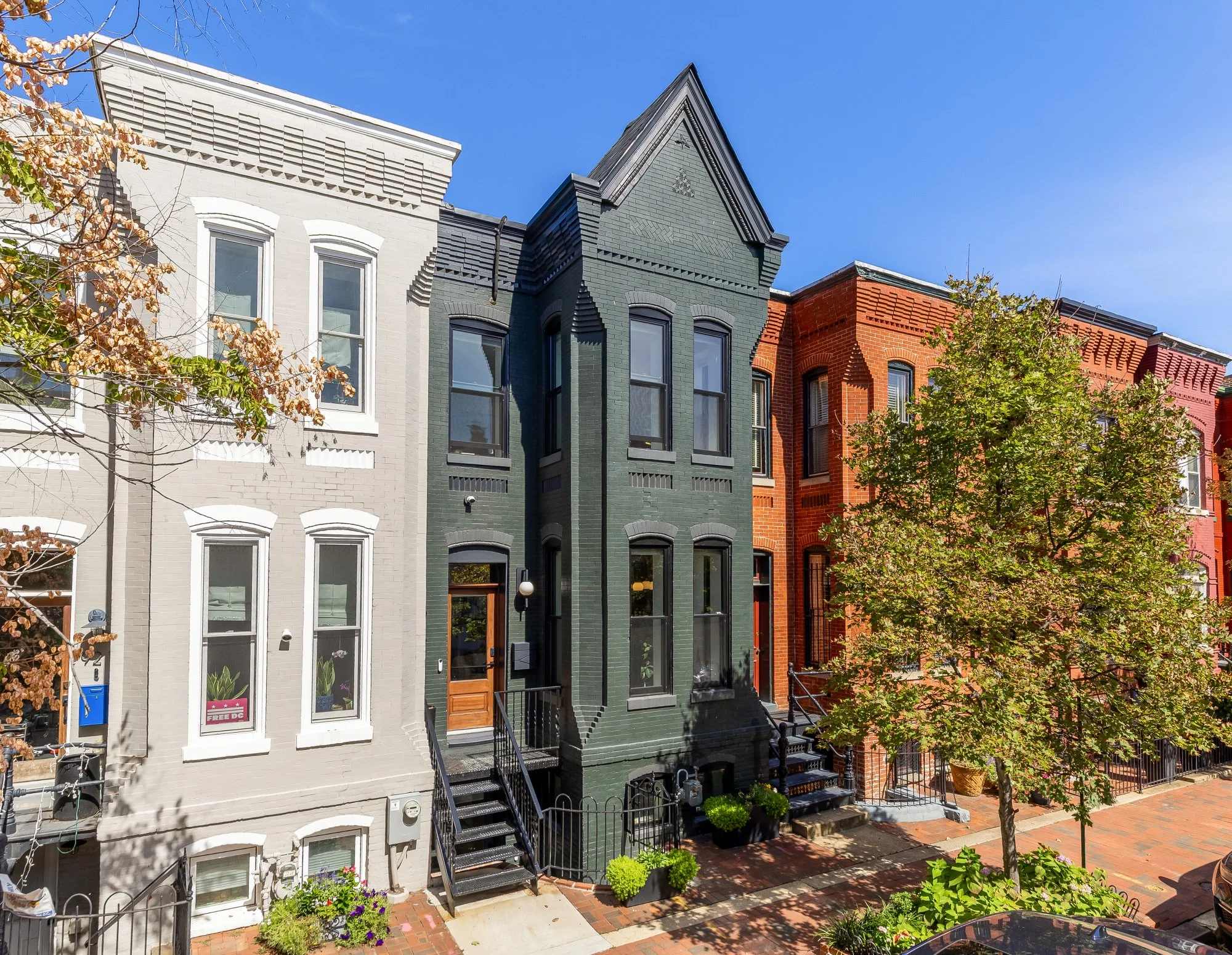 Row of colorful attached townhouses with black staircases leading to wooden front doors, green trees and brick sidewalk in front, and clear blue sky above.