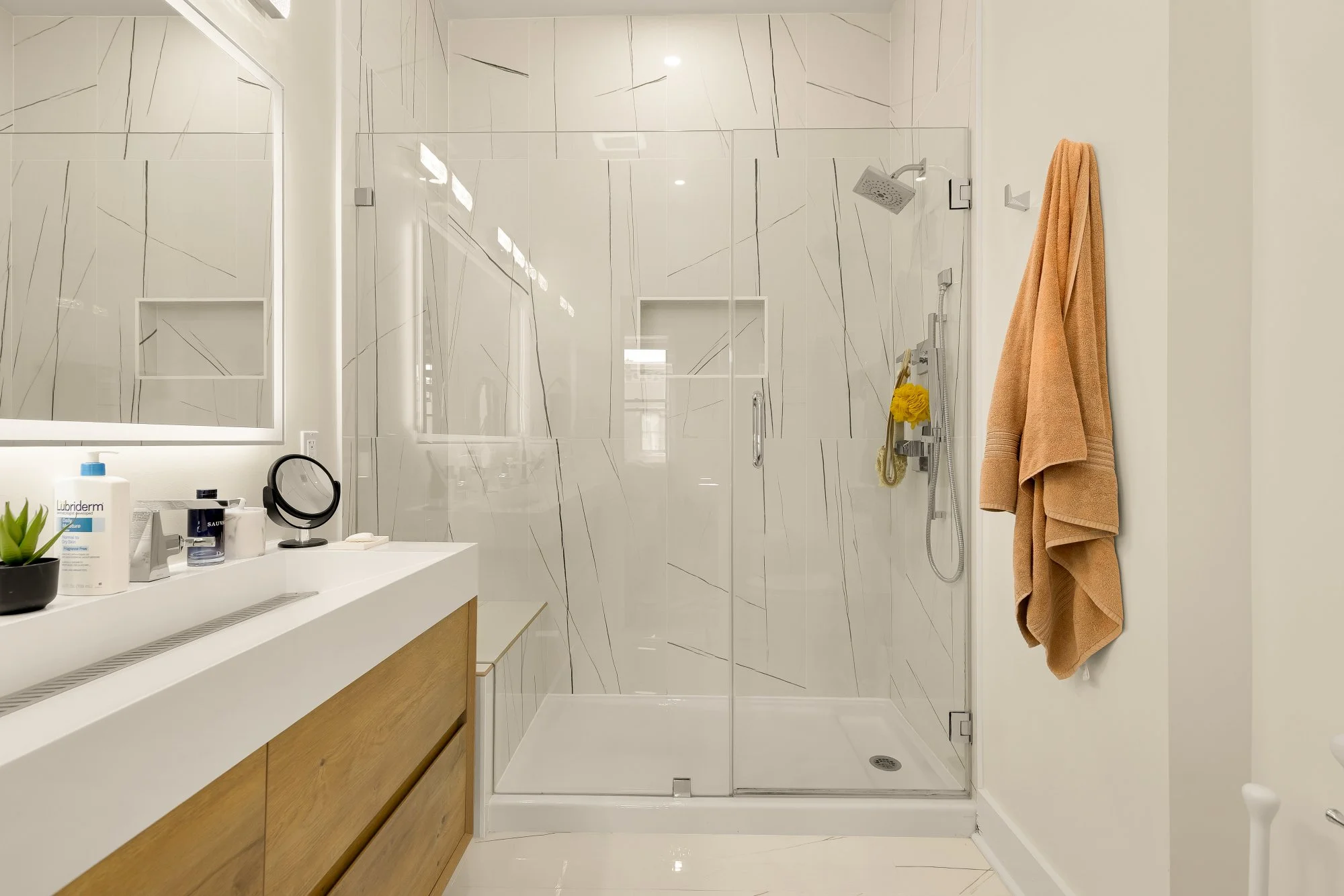 Modern bathroom with a walk-in shower with glass door, a beige towel hanging on a hook, a wooden vanity with a white countertop, and various toiletries on the counter. The walls and shower are decorated with large white tiles with gray veining.