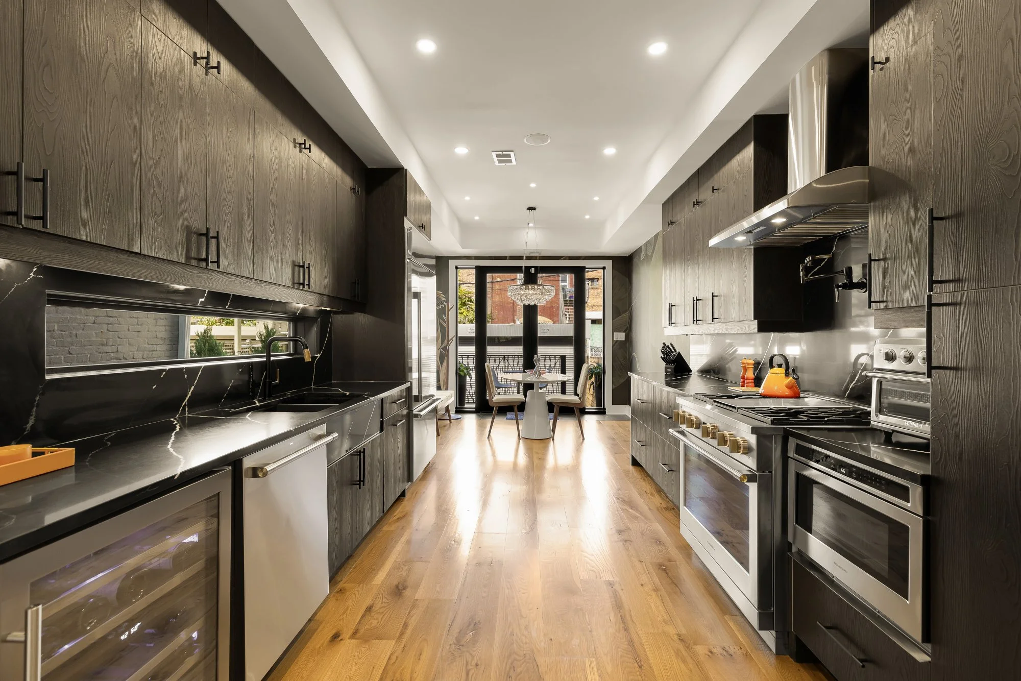 Modern kitchen with dark wood cabinets, black marble countertops, stainless steel appliances, and hardwood flooring. Small dining table with four chairs and a large window at the back.