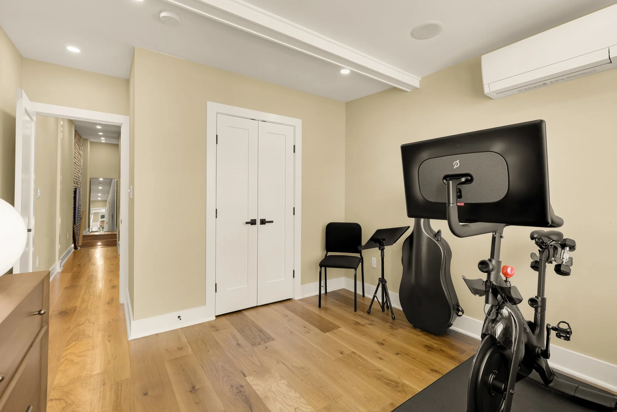A home gym with exercise equipment including a stationary bike and a treadmill, a black chair, and a wall-mounted television. The room has beige walls, white trim, a wooden floor, and an air conditioning unit on the wall.