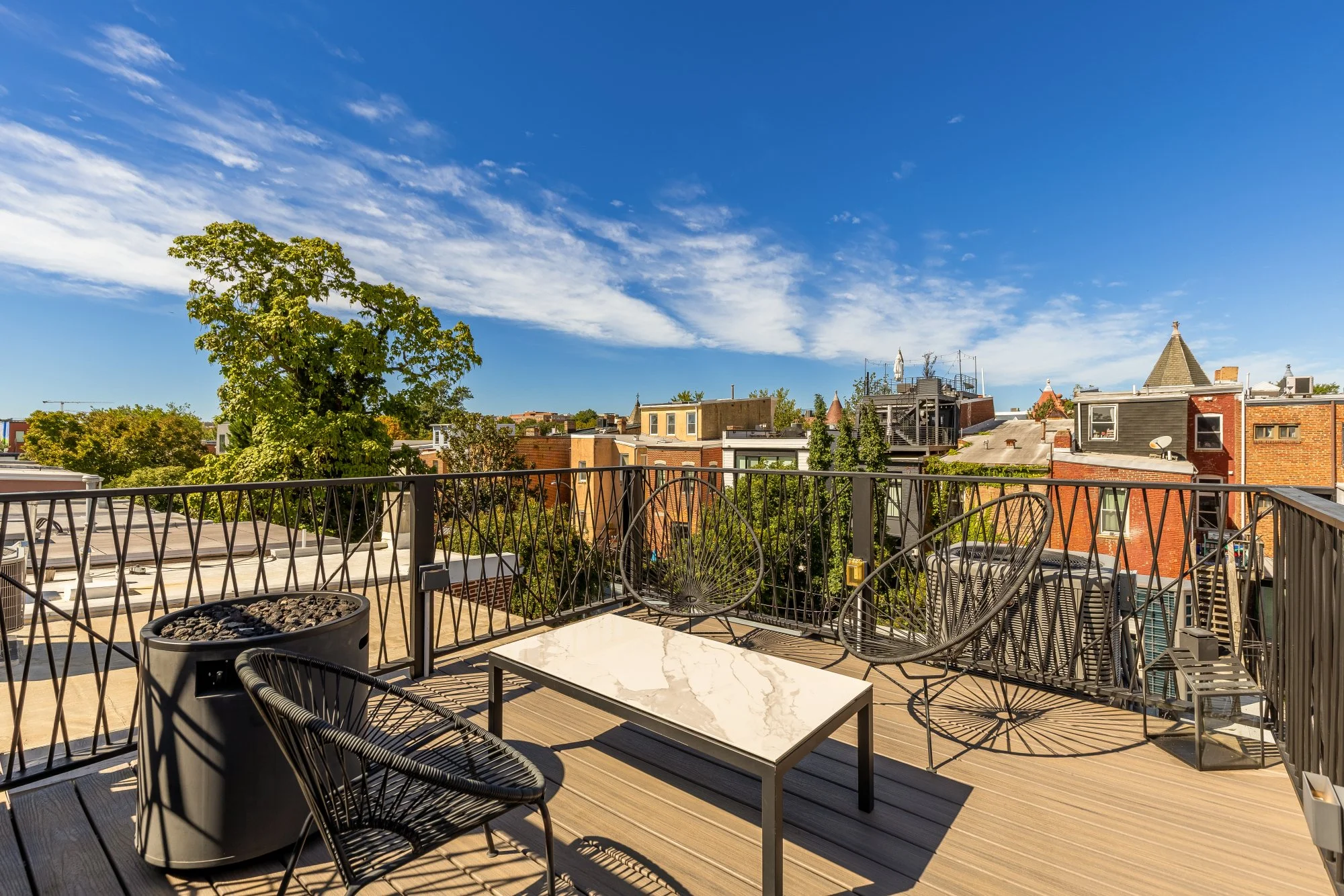 Balcony with black metal chairs and a white marble table, overlooking rooftops and a tree under a blue sky with scattered clouds.