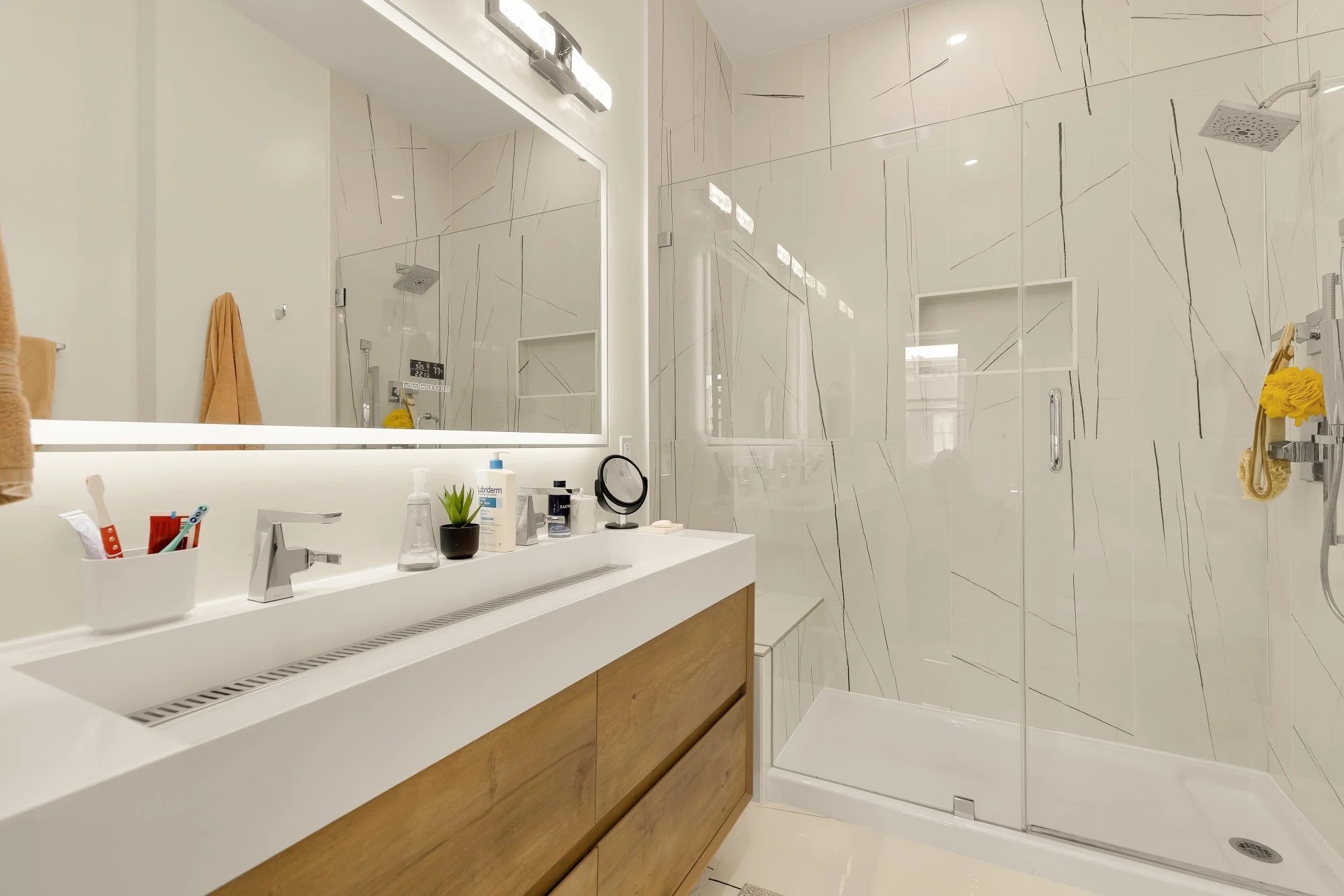 Modern bathroom with white marble walls, a white countertop with a small plant, toothbrush holder, and toiletries, wooden cabinet, large mirror, and walk-in shower with glass door and rain showerhead.