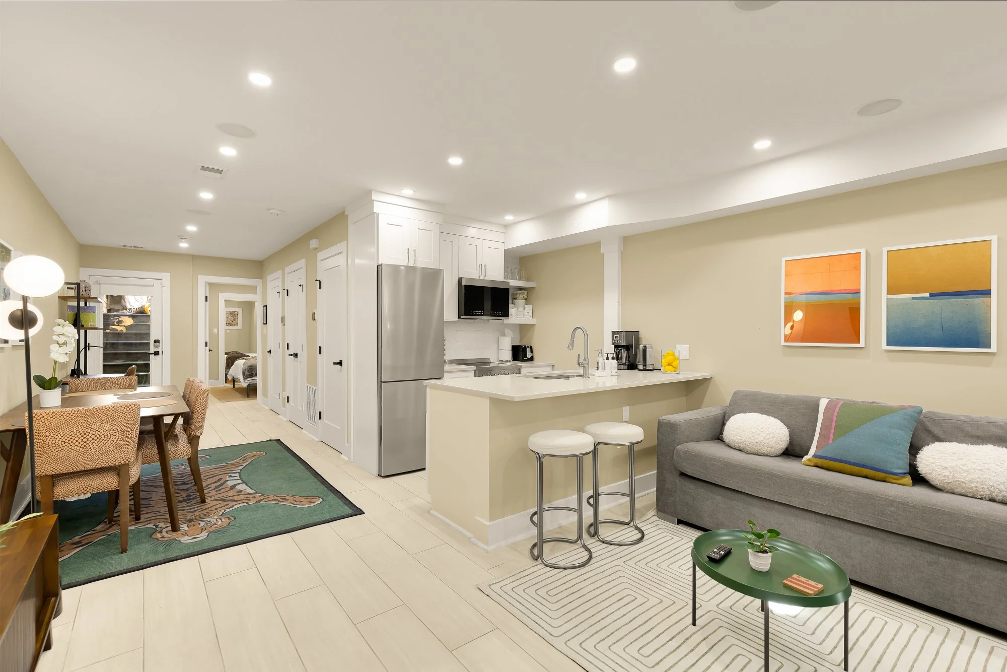 Open-concept living area with beige walls, white kitchen cabinets, stainless steel appliances, a kitchen island with stools, a gray sofa with colorful pillows, a dining table with chairs, and artwork on the wall.