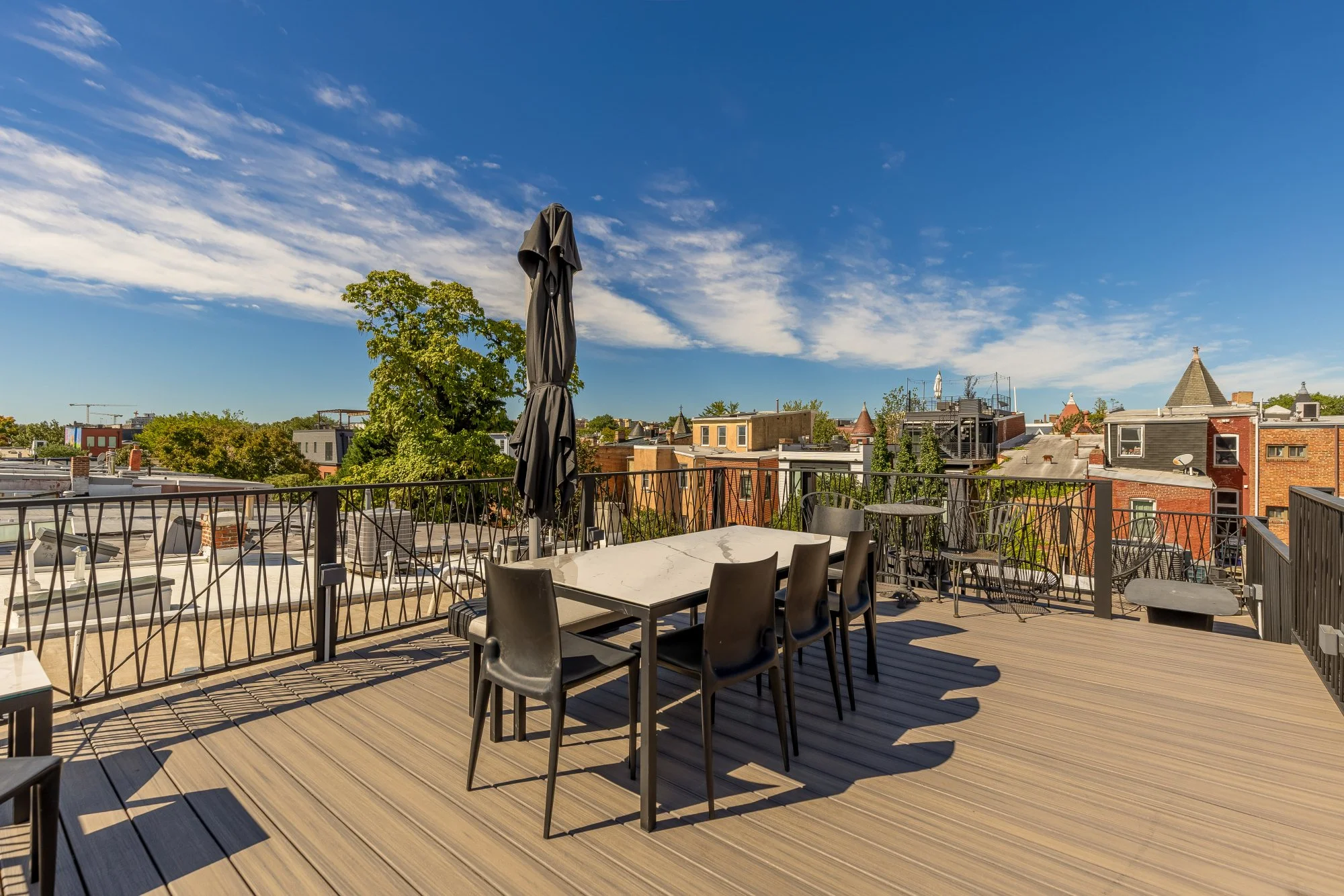 A spacious rooftop patio with wooden decking, black chairs around tables, and a closed black umbrella, overlooking a cityscape with trees and rooftops under a clear blue sky with scattered clouds.