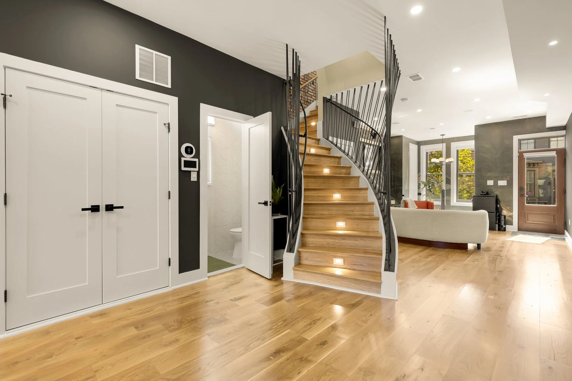 Modern interior of a house with wooden flooring, a staircase with black metal railing, and large windows allowing natural light. There is a white sofa, a small plant, and an open door leading to a bathroom.