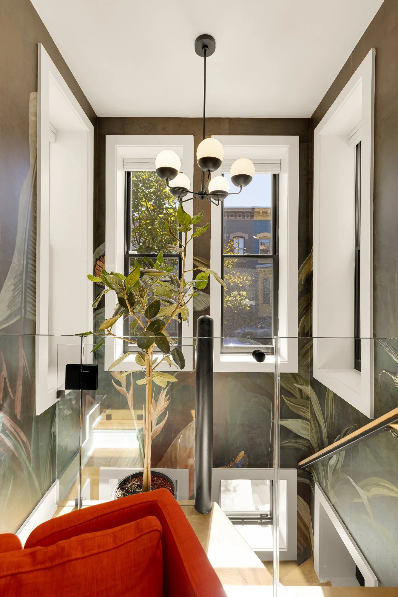 Interior view of a staircase landing with large windows, a potted plant, a modern chandelier, a red upholstered chair, and decorative wall panels with botanical patterns.