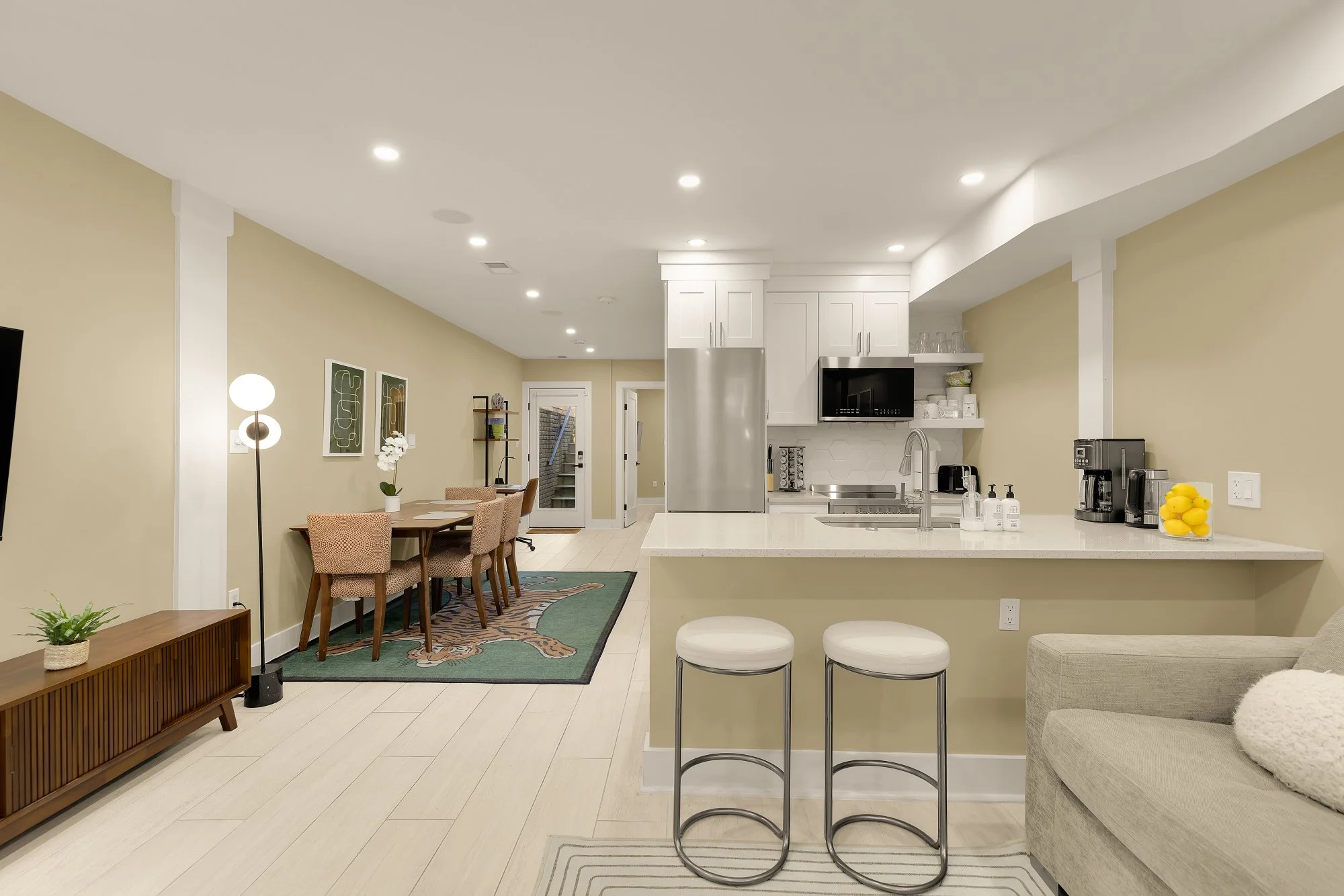 Open-concept kitchen and dining area with beige walls, white cabinetry, stainless steel appliances, and a breakfast bar with white barstools. Leather armchair in the foreground, dining table with upholstered chairs, and contemporary artwork on the wa