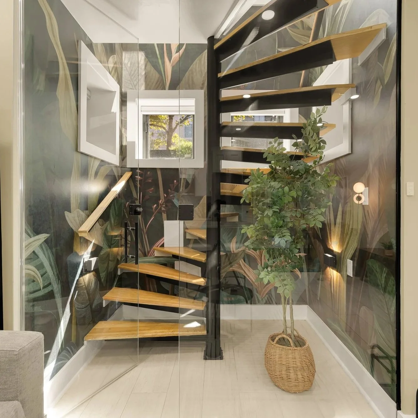 Interior view of a modern stairway with wooden steps and black metal support, surrounded by leafy green plants, and background wall adorned with large botanical wallpaper.