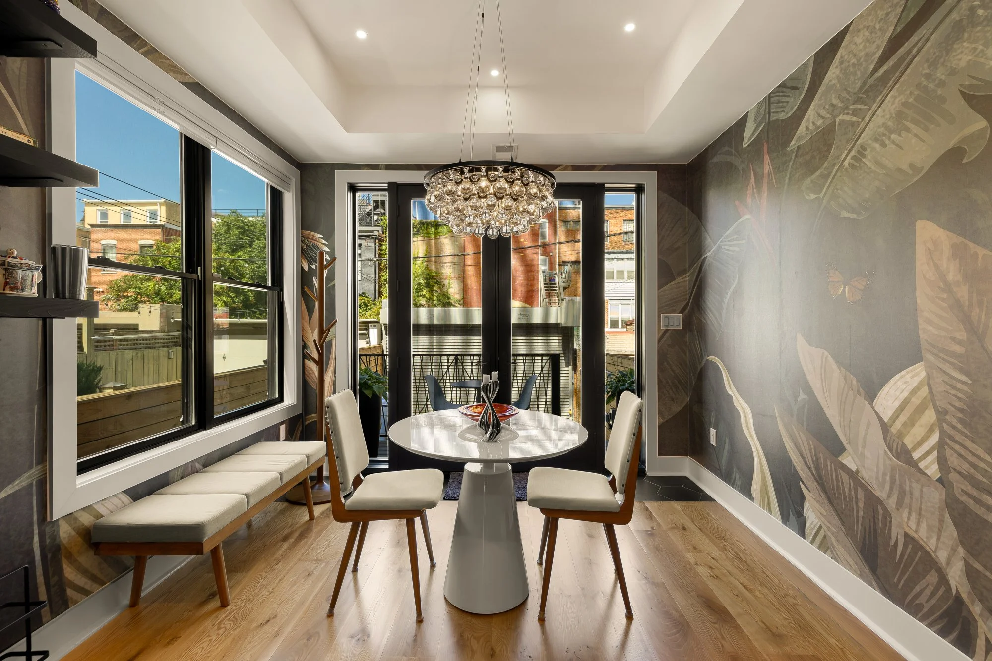 Interior view of a modern dining room with large windows, a round table with white chairs, a built-in bench along the wall, a chandelier hanging above the table, and a decorative wall mural with tropical leaves. Outside, a balcony with railing and ne