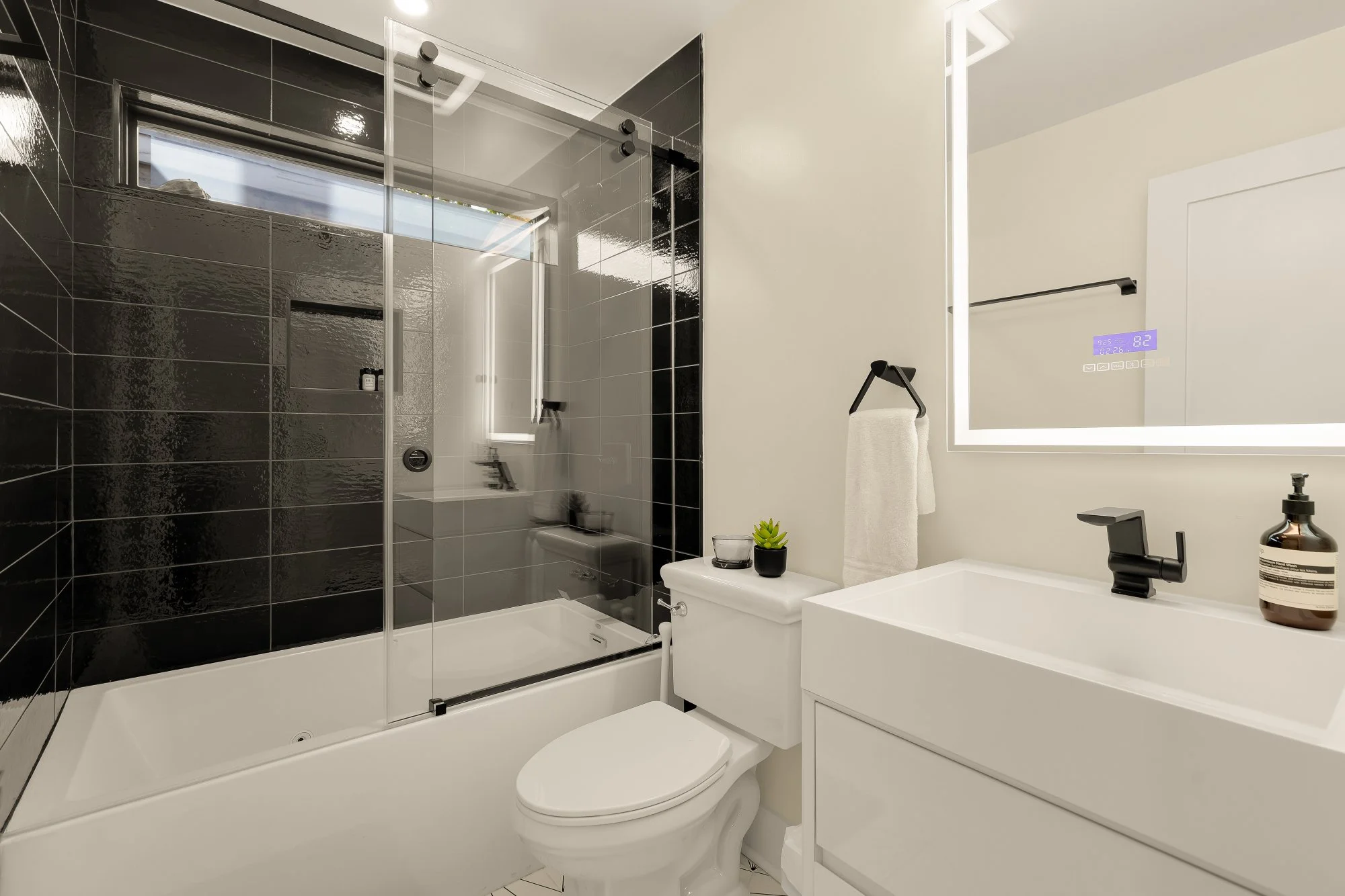 Modern bathroom with a black-tiled shower area with glass sliding doors, a small window, and a white vanity with a large illuminated mirror, a towel hanging on a black towel bar, a small potted plant, and a bottle of soap or lotion.