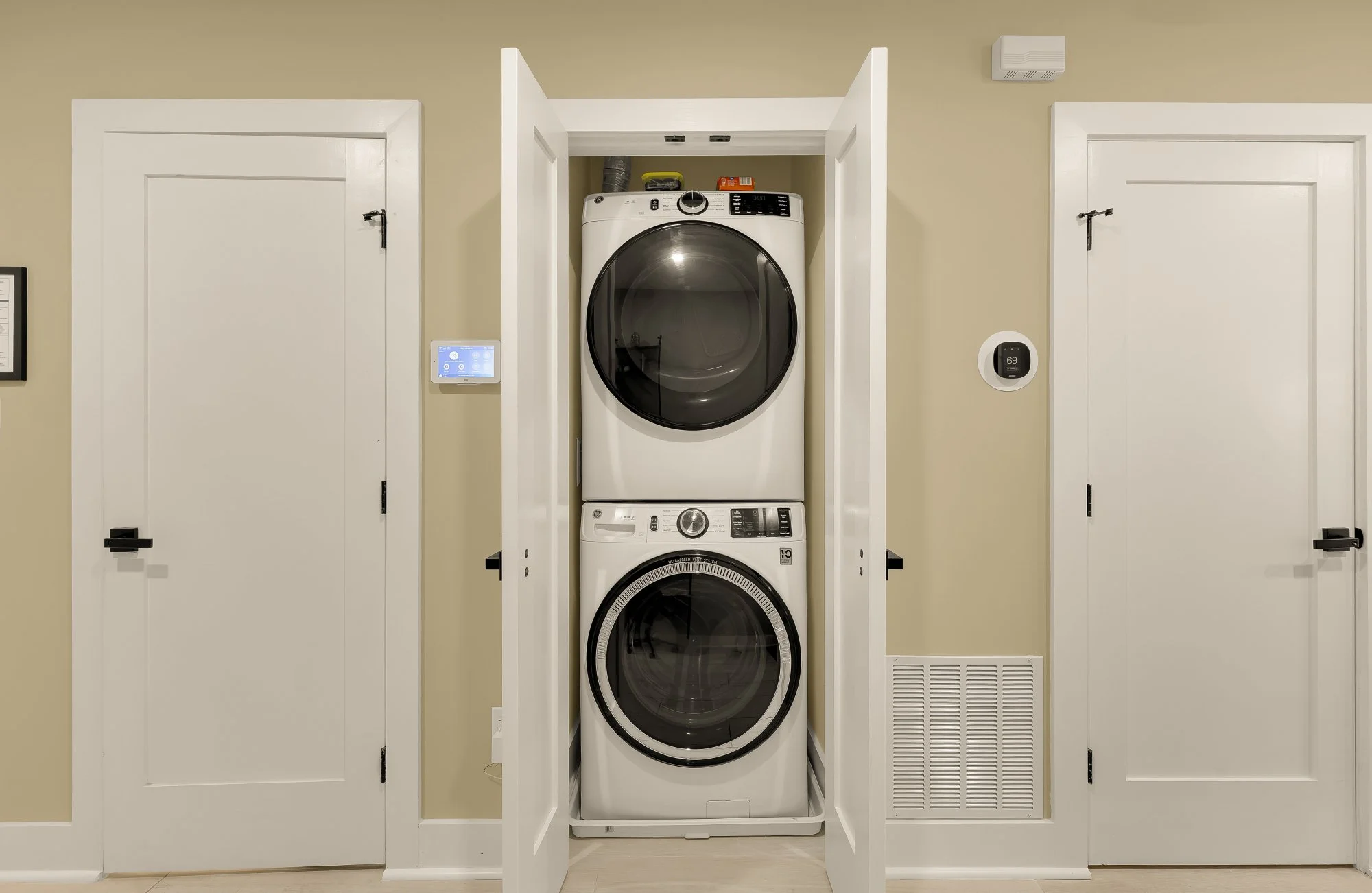 Stacked washer and dryer in a closet with doors open, flanked by two closed doors, digital thermostat, and ventilation.