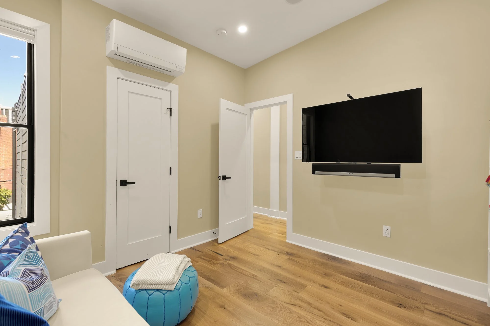 A living room with a beige wall, a flat-screen TV mounted on the wall, a white door, a window with sunlight, a white sofa with patterned pillows, and a blue ottoman.
