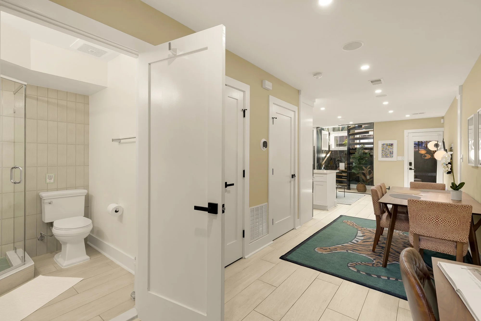 Interior of a modern home with a view of a living area, dining table, and a bathroom with a toilet and shower. The space is decorated with plants and artwork.