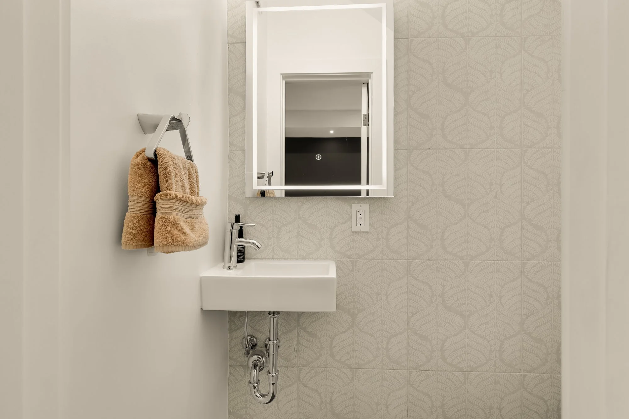 Bathroom sink with beige towels hanging on a wall-mounted towel rack, a mirror, and a small sink with a chrome faucet. Part of the bathroom wall with decorative tiles is visible.