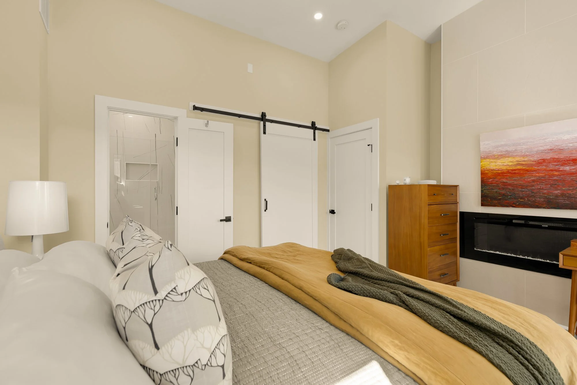 Interior view of a bedroom with a bed, pillows, a blanket, a nightstand with a lamp, a wooden dresser, a colorful landscape painting on the wall, and a black electric fireplace.