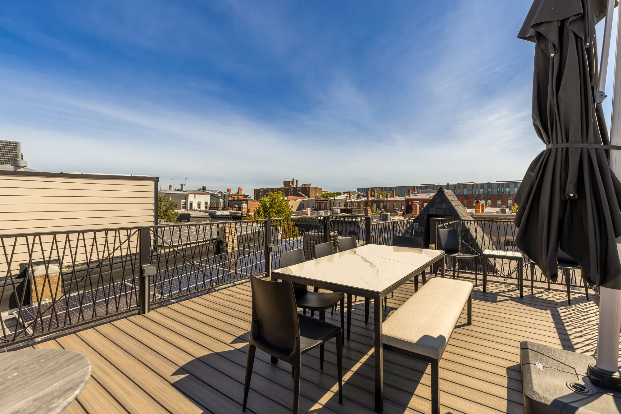 A rooftop outdoor patio with seating, tables, and an umbrella, overlooking a cityscape under a partly cloudy sky.