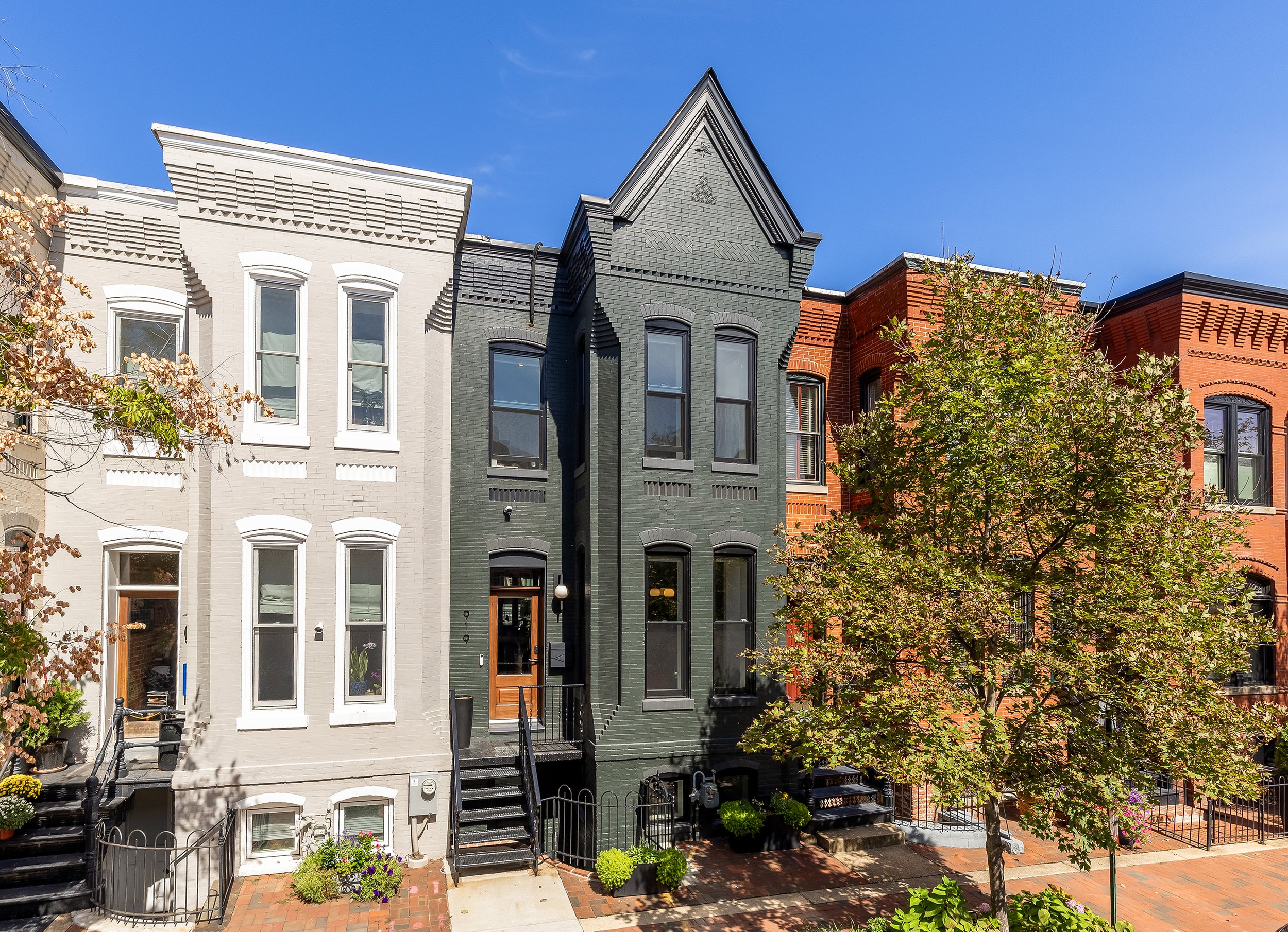 Colorful row of historic townhouses with front stairs, brick facades in white, gray, and red, and trees in the foreground under a clear blue sky.