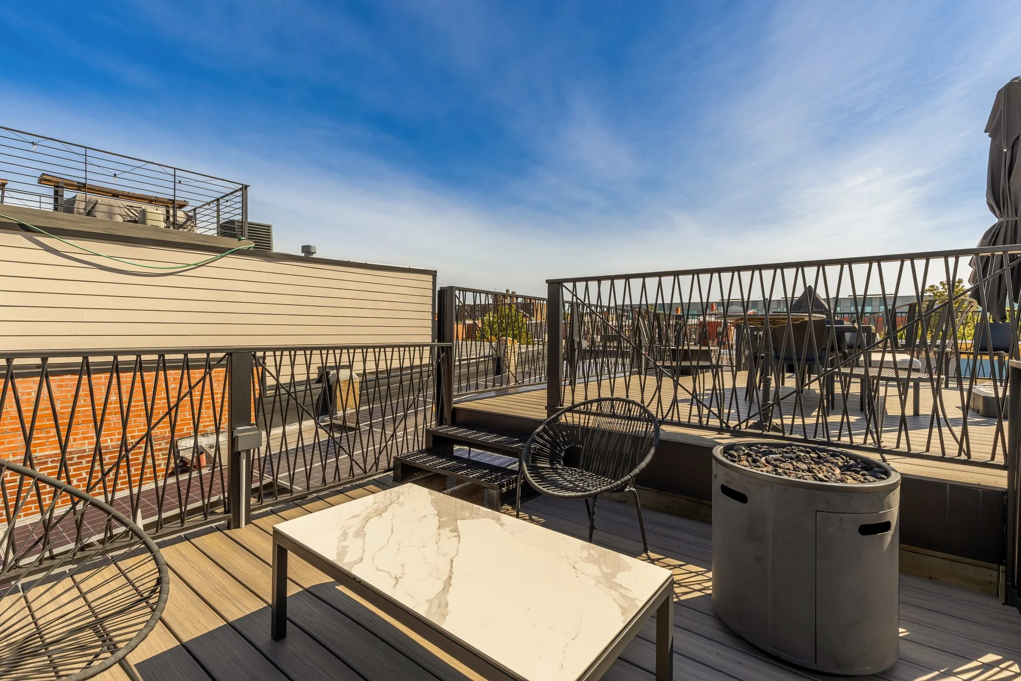 A rooftop terrace with modern outdoor furniture, including a marble-top coffee table and black chairs, surrounded by a metal railing. There are some chairs and planters, with a clear blue sky overhead.