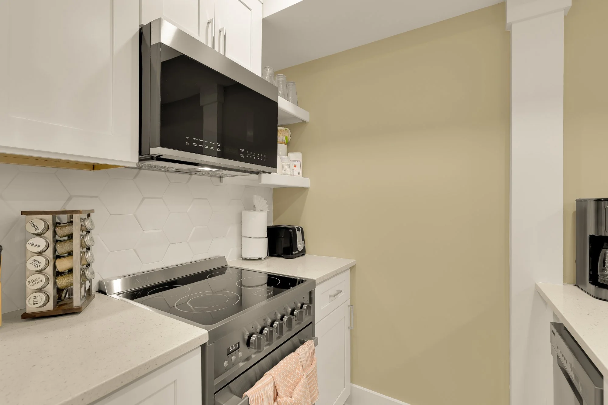 A modern kitchen with white cabinets, a black microwave, and a stainless steel stove with an oven. There are spice jars on a wooden rack, a paper towel roll, and small appliances like a toaster and a coffee maker on the white countertop. The walls ar