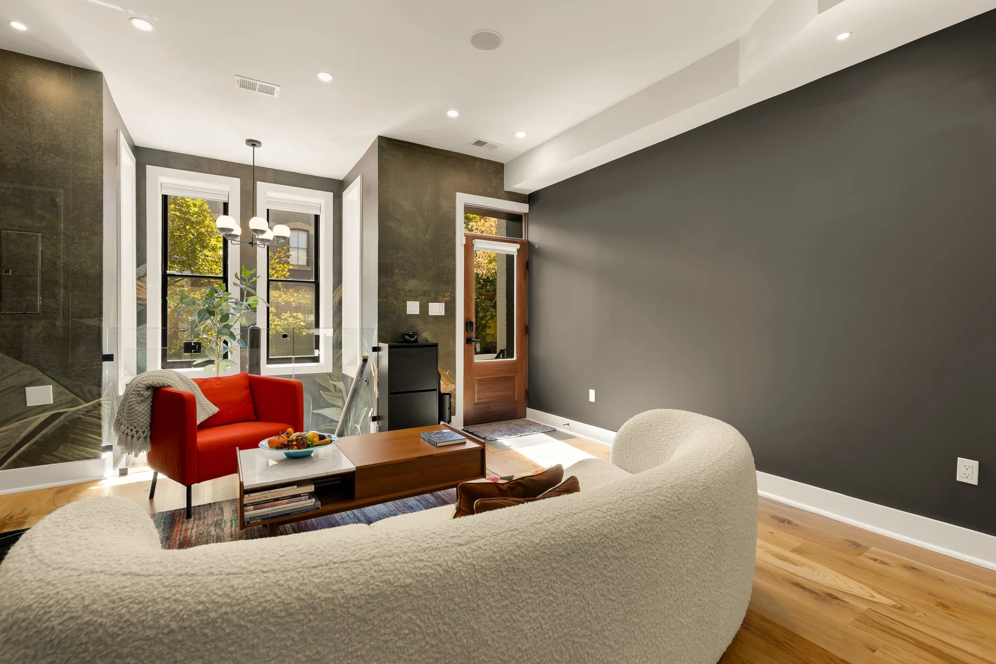 Living room with large dark gray wall, beige sofa, red armchair with blanket, wooden coffee table with fruit bowl, books underneath, and houseplants near windows with leaves visible outside.