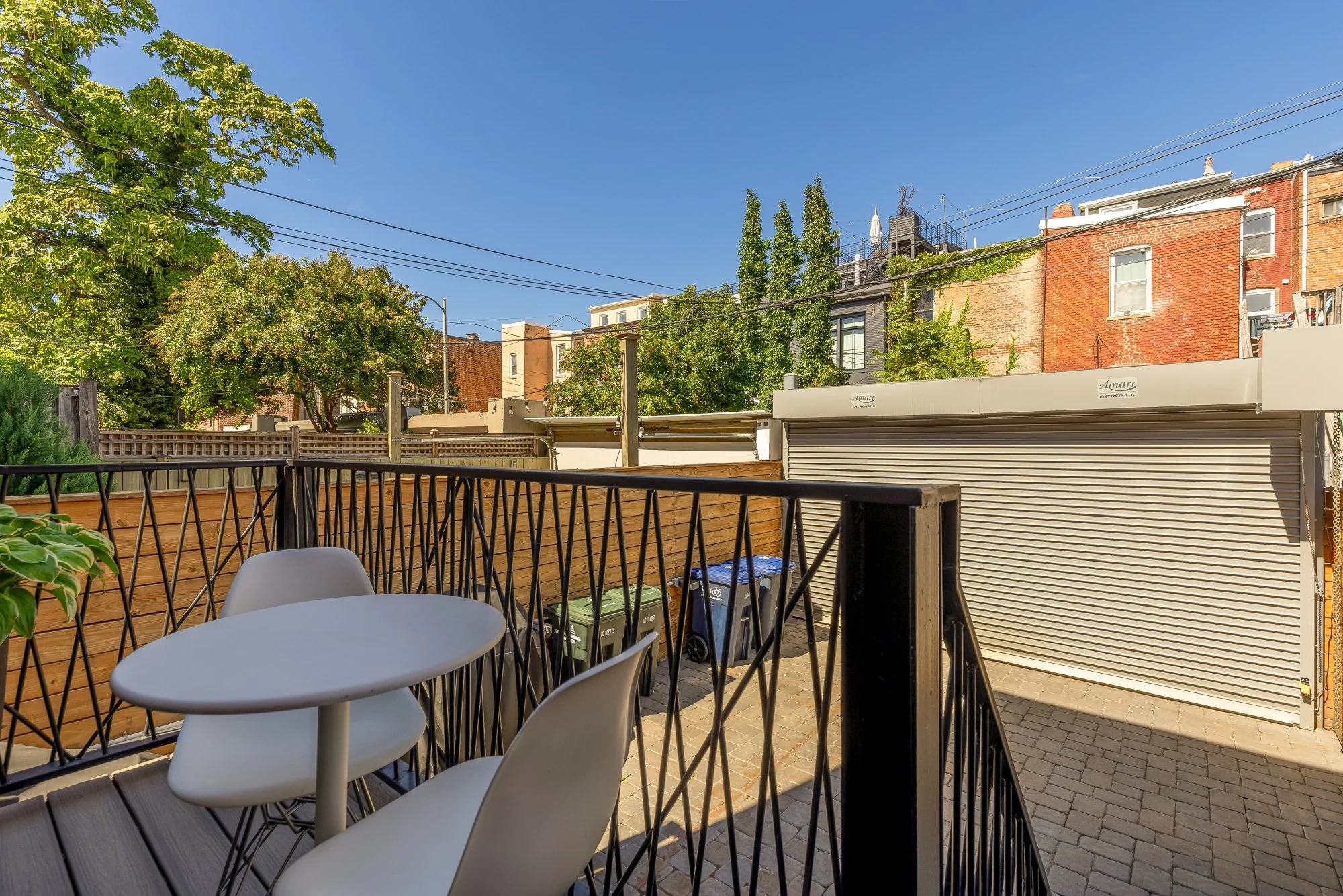 Backyard patio with white chairs around a small white table, wooden fence, and trees, with buildings and utility wires in the background under a clear blue sky.