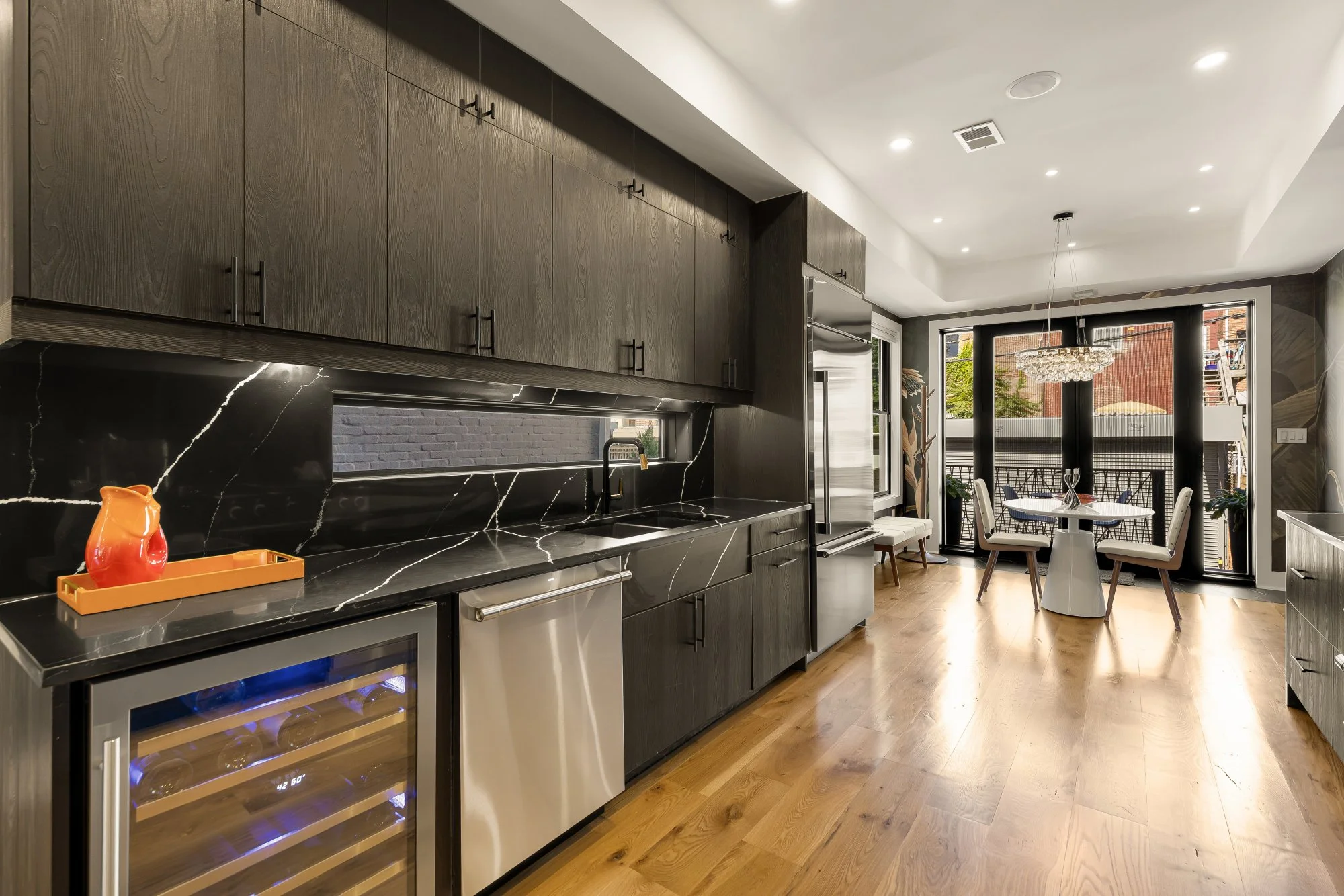 Modern kitchen with dark wood cabinets, marble backsplash, stainless steel appliances, a small wine fridge, and a dining area with a round table and chairs near large windows and a balcony.