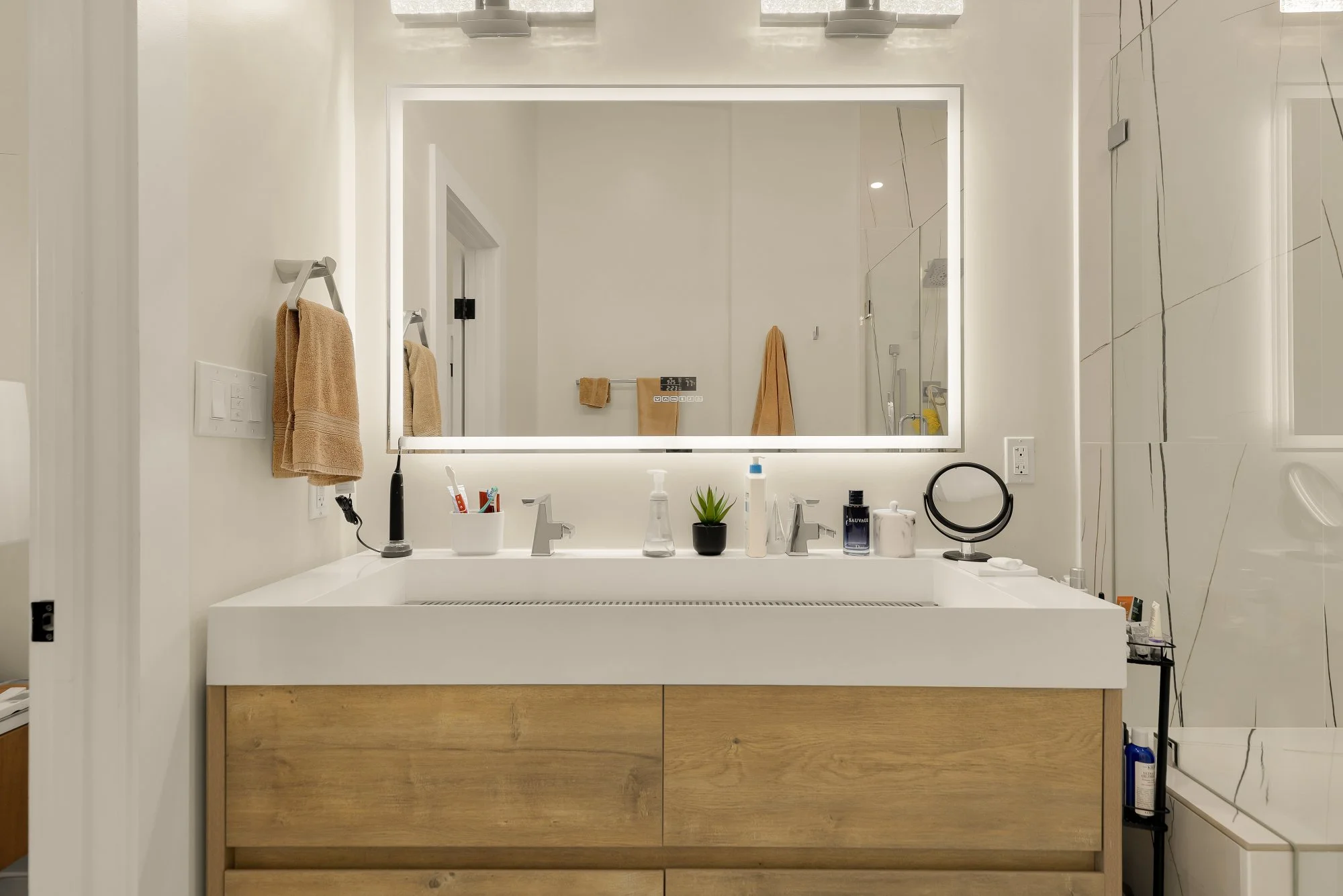 Modern bathroom vanity with a large illuminated mirror, beige towels hanging on hooks, toothbrushes, and toiletries on the countertop, and a white and marble wall.