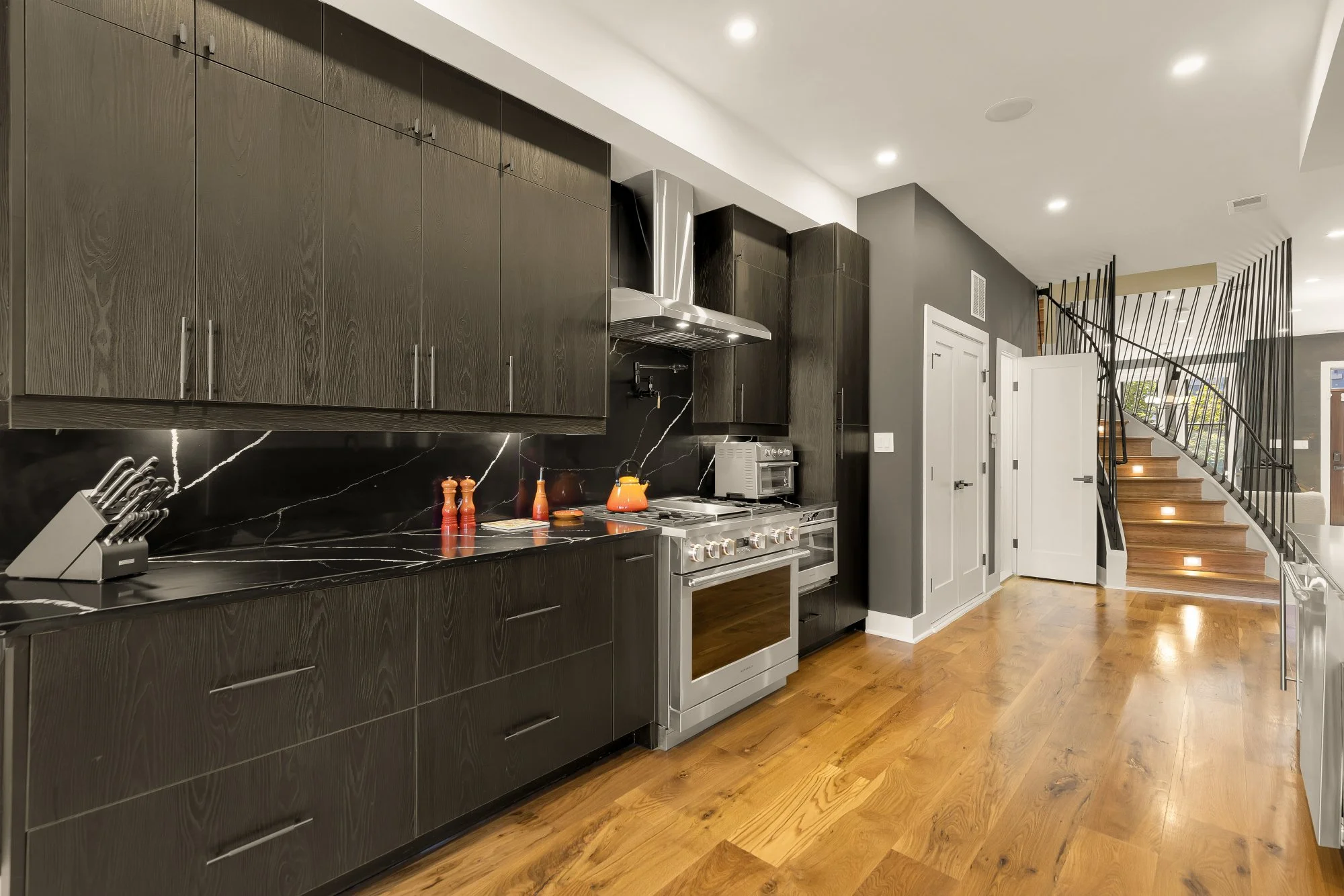 Modern kitchen with dark wood cabinets, black marble countertops, stainless steel oven and range, and a staircase with wooden steps and black metal railing.