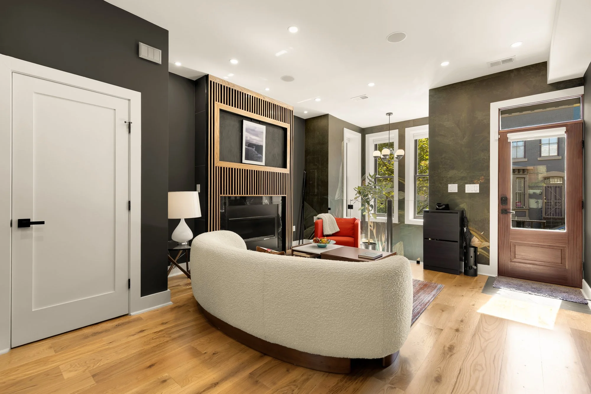 Interior view of a modern living room with wooden flooring, white and dark gray walls, large windows, a white upholstered sofa, a small dark wooden table, a red armchair, a floor lamp, and decorative elements including framed pictures and greenery.