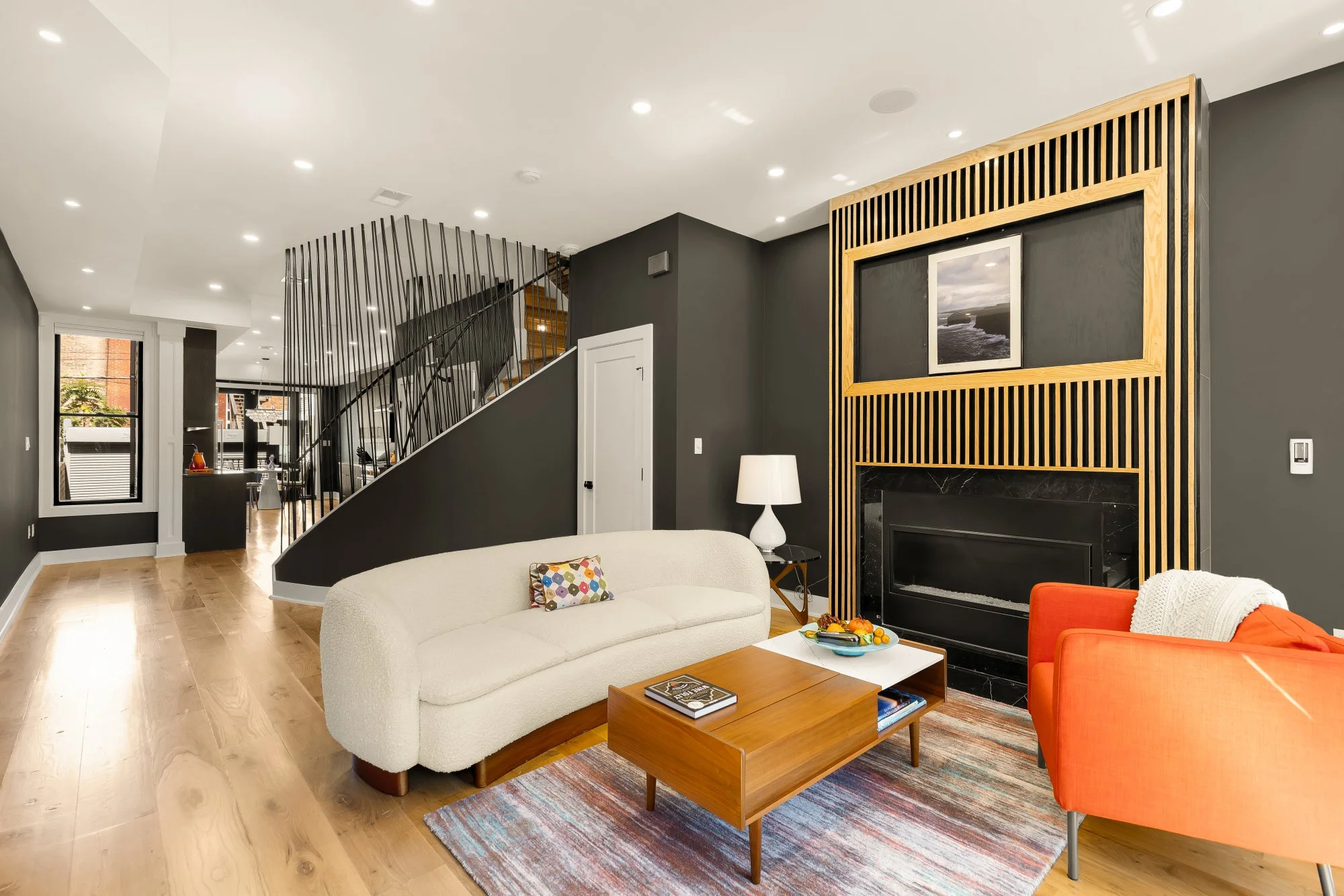 Modern living room with white and dark gray walls, a cream sofa, orange armchair, coffee table with books and snacks, fireplace with wooden frame, and a staircase with black metal railing.