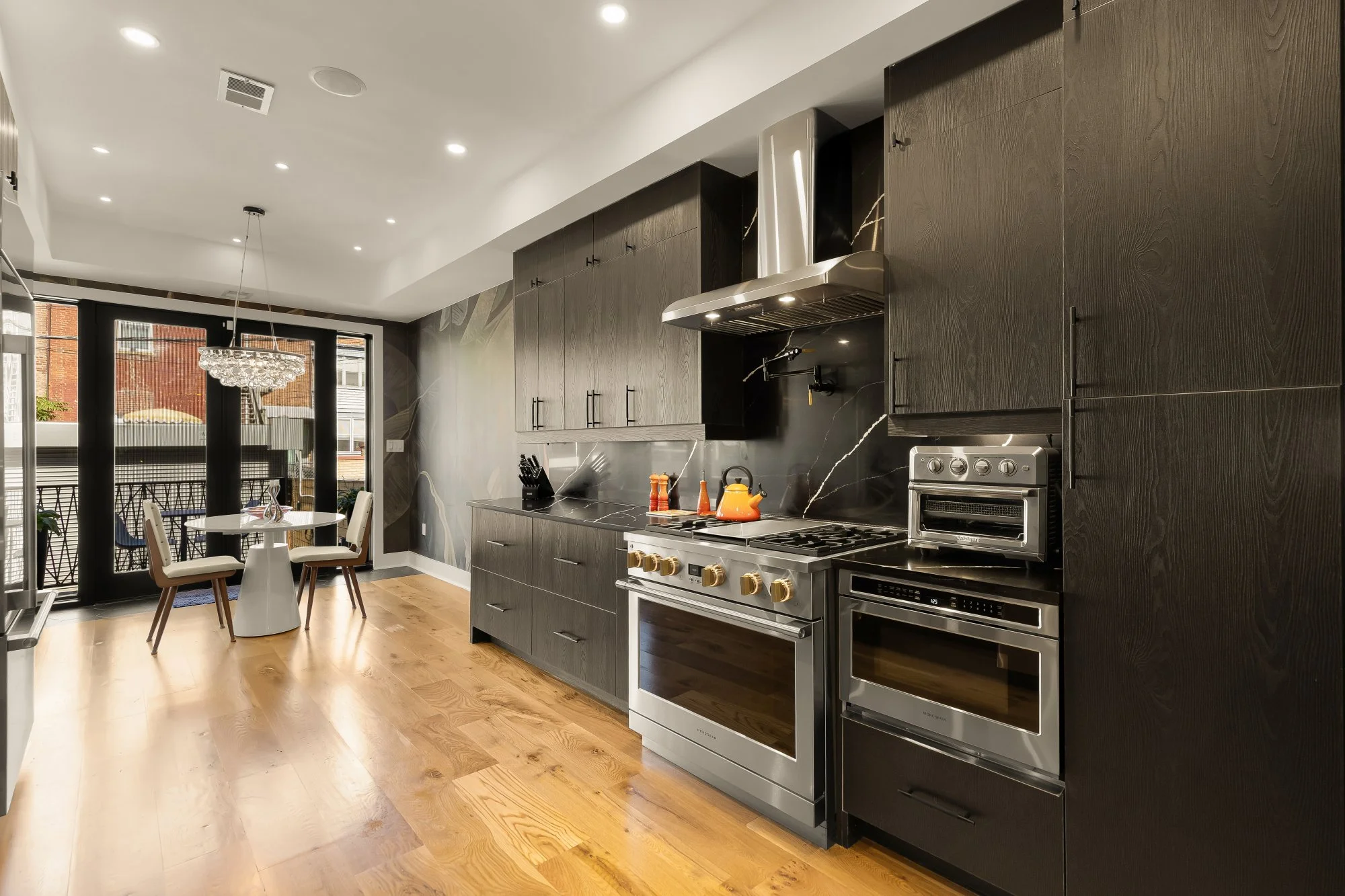 Modern kitchen with black cabinetry, stainless steel appliances, and hardwood flooring. There is a small dining area with a white table and four chairs, and a balcony with outdoor furniture outside the glass doors.