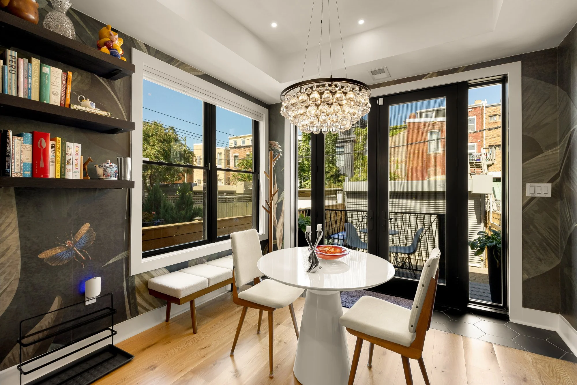 A modern dining room with a white round table surrounded by four white chairs, a built-in window seat with cushions, and a large sliding glass door leading to a small balcony. The room features dark patterned walls, a chandelier with glass globes, an