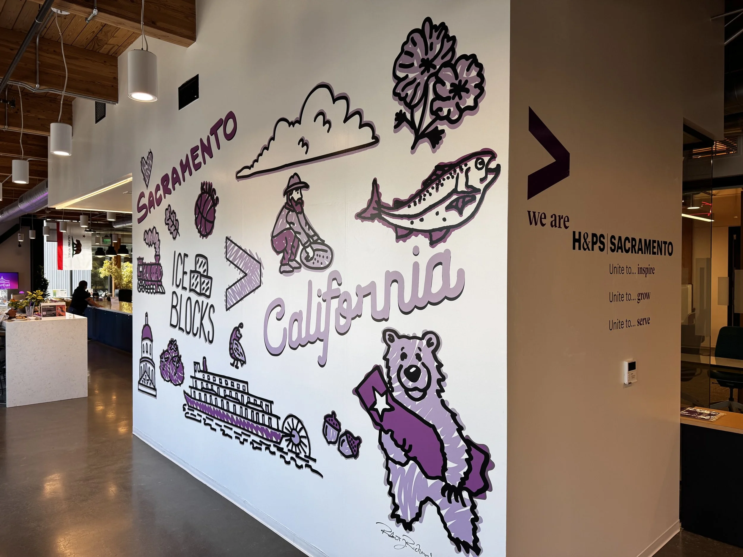 Interior of a modern office or community space with a large wall illustration featuring California-themed drawings, including a bear, a fisherman, a mountain, a train, and related text and symbols.