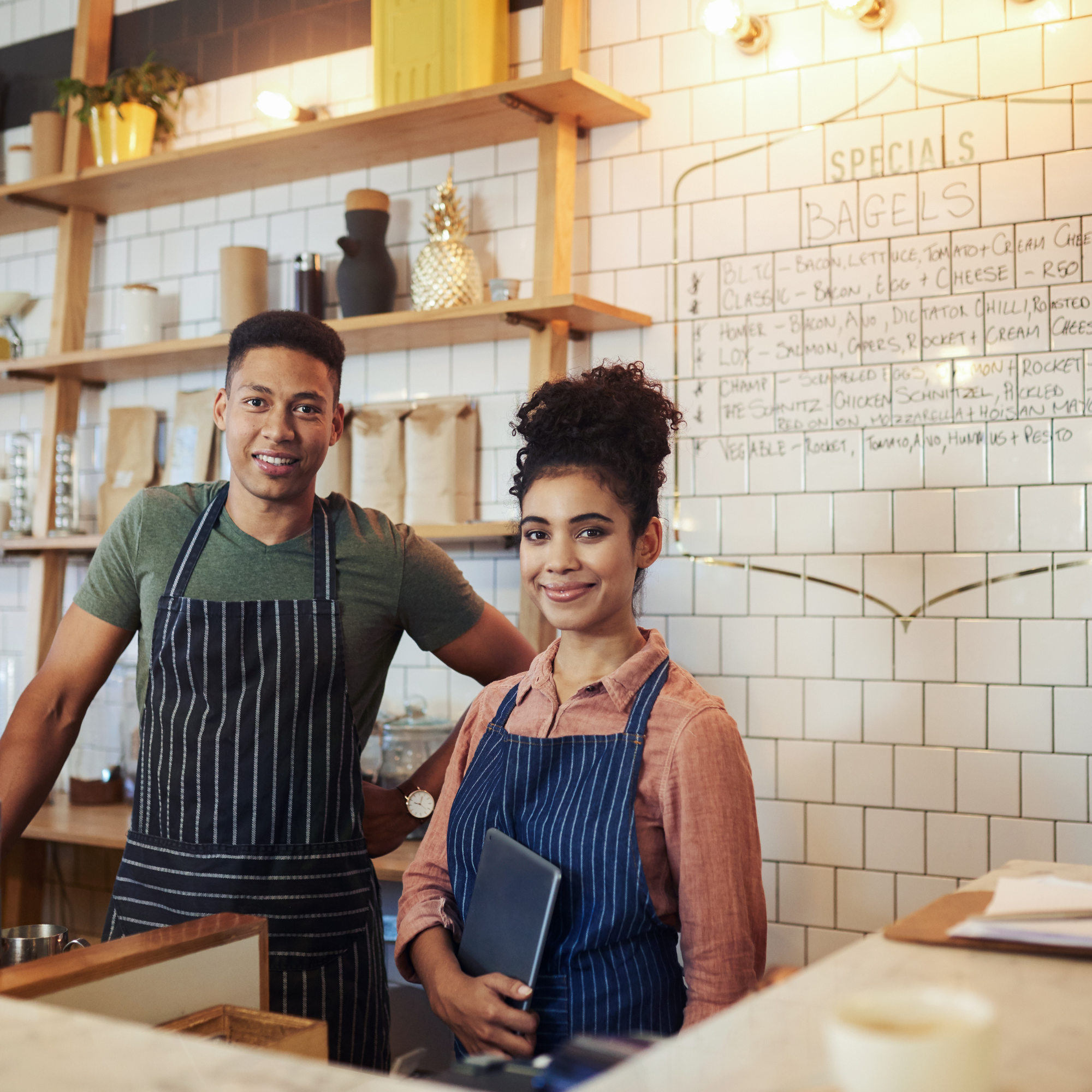 Two waitstaff women in aprons standing in a restaurant kitchen area with a white subway tile wall and shelves decorated with potted plants, a decorative pineapple, and bottles. One woman holds a tablet and smiles, the other faces away from the camera, both wearing casual clothing and aprons. A menu board with handwritten items is visible in the background.