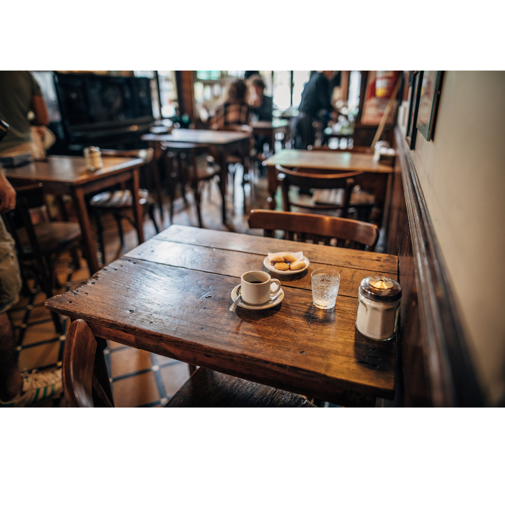 Interior of a cozy café with wooden tables and chairs, a coffee cup, a glass of water, a small plate with cookies, and various decorations on the walls, with blurred patrons in the background.