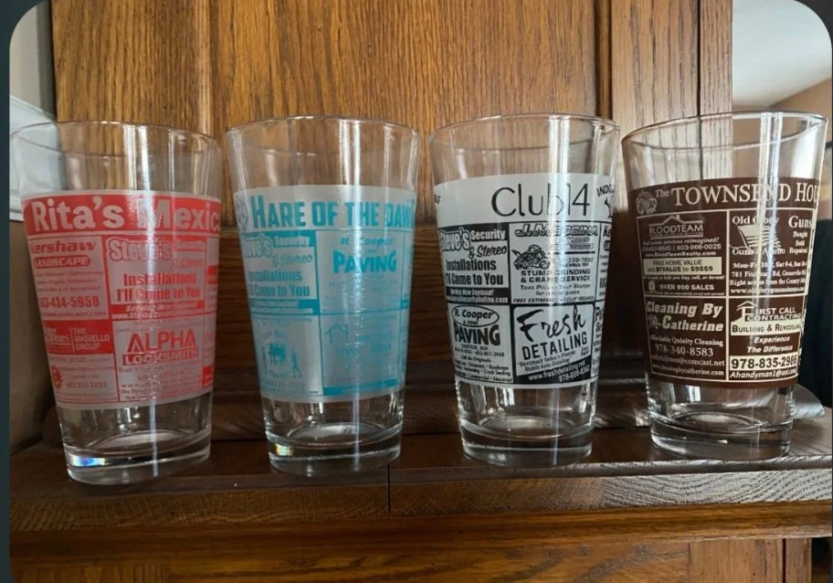 A row of five clear drinking glasses with various company logos and advertisements on them, placed on a wooden shelf.