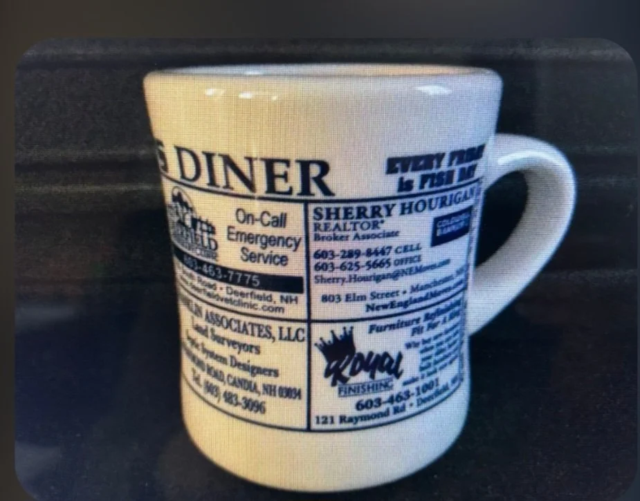 A ceramic coffee mug with printed advertisements and business information, including emergency services, real estate, and furniture.