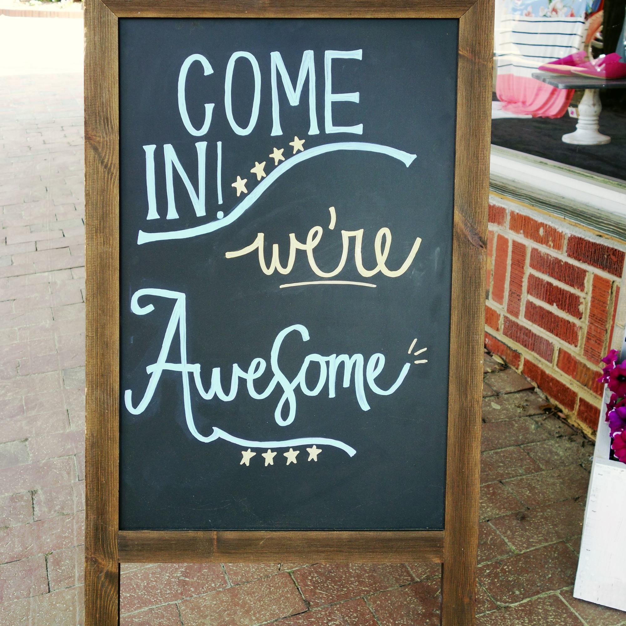 A black chalkboard sign with a wooden frame displaying the message 'Come in! We're awesome!' in white and gold handwritten text, decorated with small stars at the bottom. The sign is placed on a brick sidewalk outside, and part of a market stall with pink shoes and striped fabric is visible to the right.
