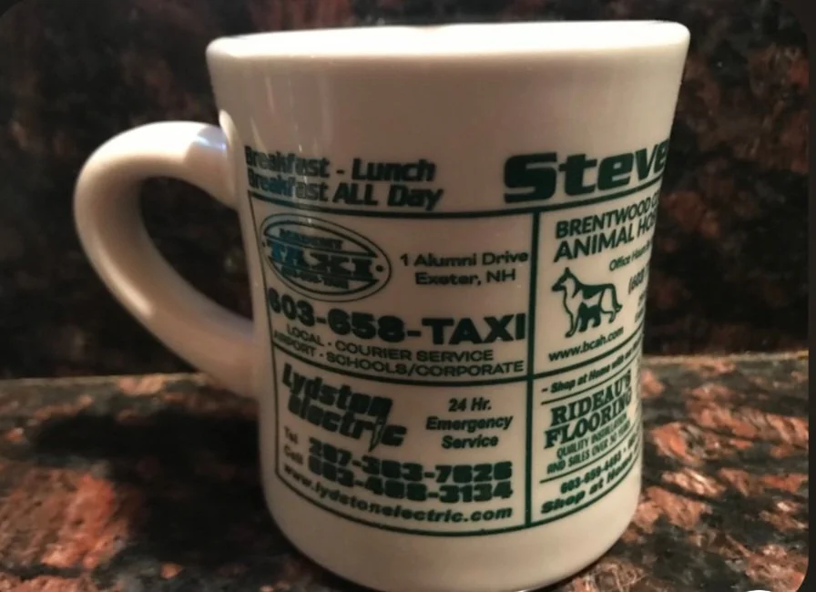 A white coffee mug with green and black printed advertisements and contact information for local businesses, including a taxi service, an animal hospital, and an electric company, placed on a dark speckled countertop.