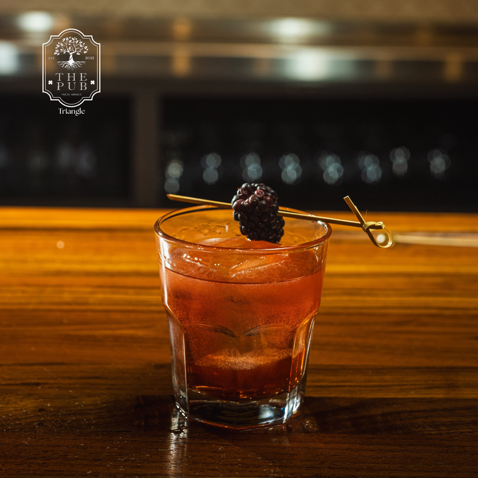 A close-up of a drink in a glass with a blackberry garnish and a cocktail pick, set on a wooden surface, with blurred background lights.