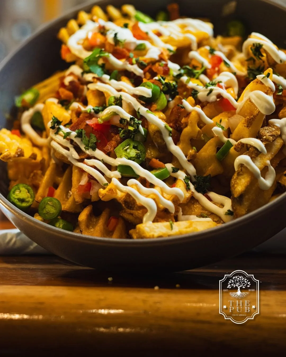A skillet of loaded nachos topped with chopped green peppers, diced tomatoes, chopped herbs, and drizzled with sour cream.