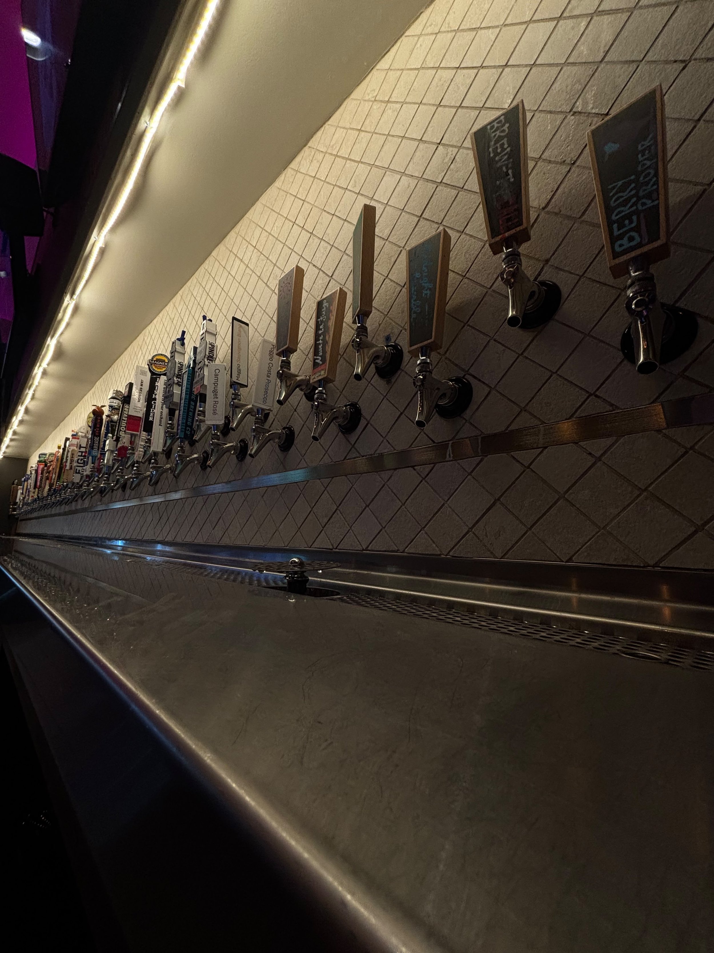 Multiple beer taps on a tiled bar wall with chalkboard signs indicating different beer options.