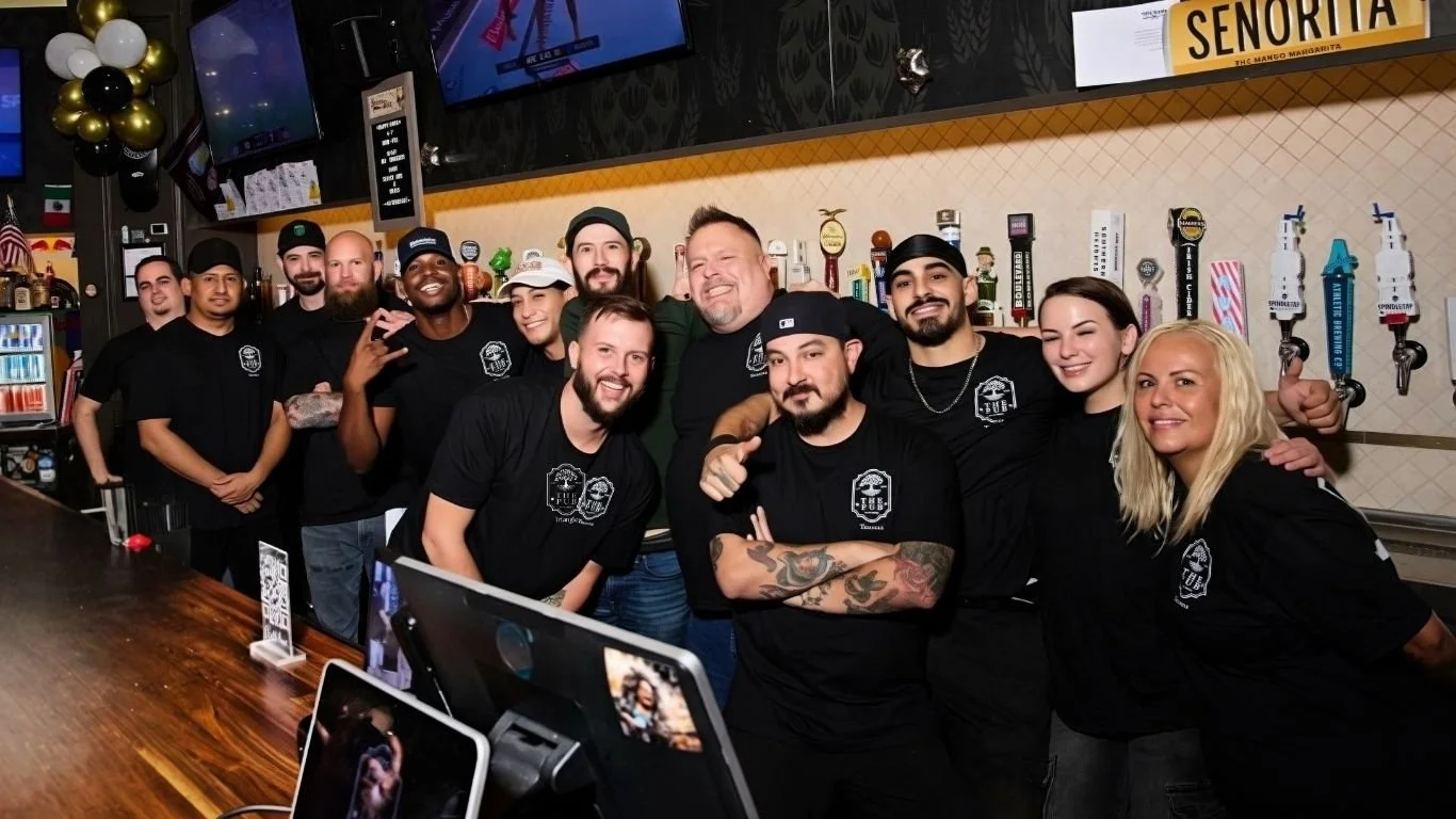 Group of people, mostly men and two women, posing together behind a bar in a restaurant or pub.