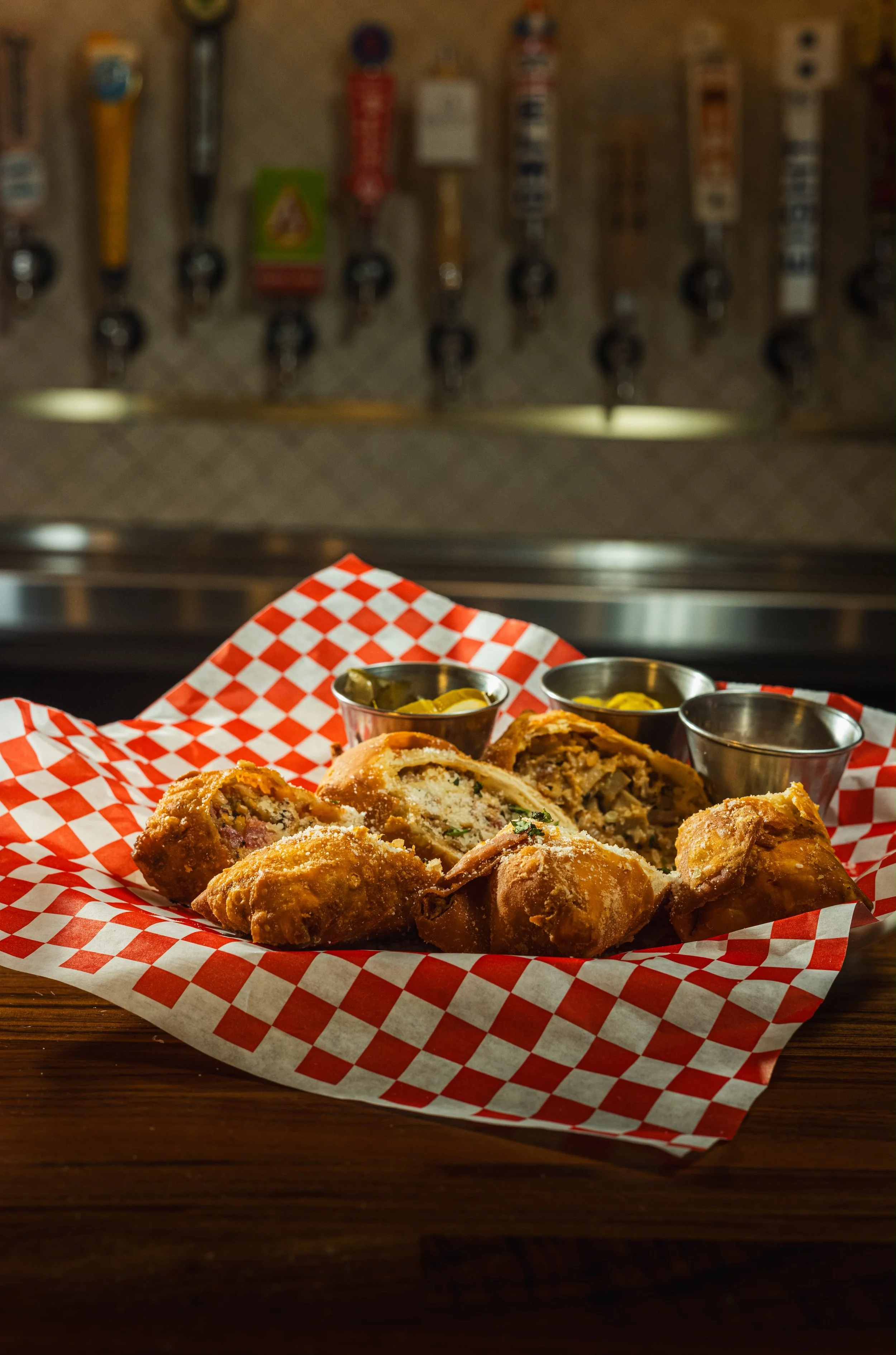 Fried food items, possibly egg rolls or spring rolls, served on a checkered paper with small cups of dipping sauces, on a wooden table, with beer taps in the background.