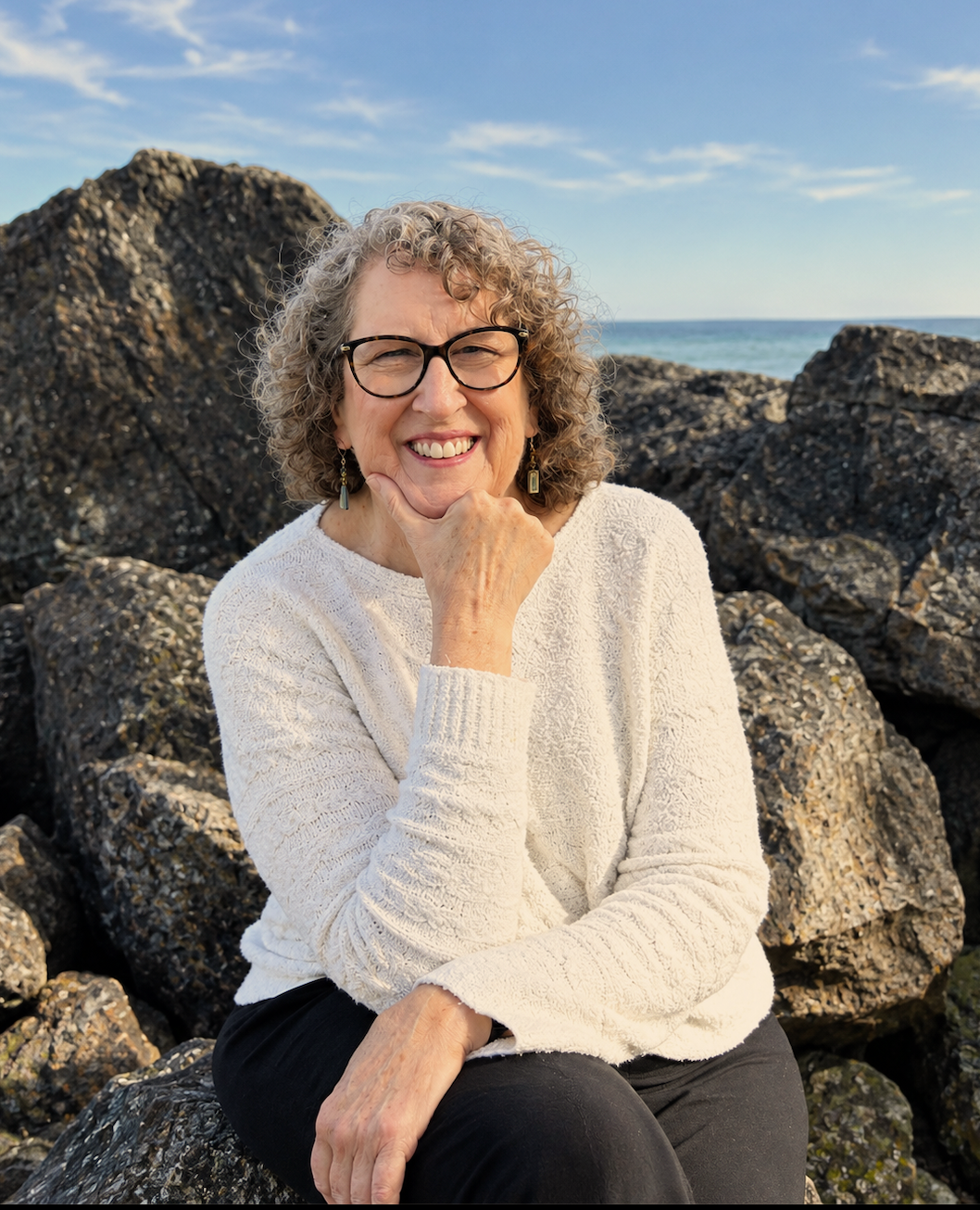 Julie Conant, Small Business Strategist. smiling woman with curly hair and glasses sitting on large rocks by the ocean, wearing a white sweater and black pants under a blue sky.