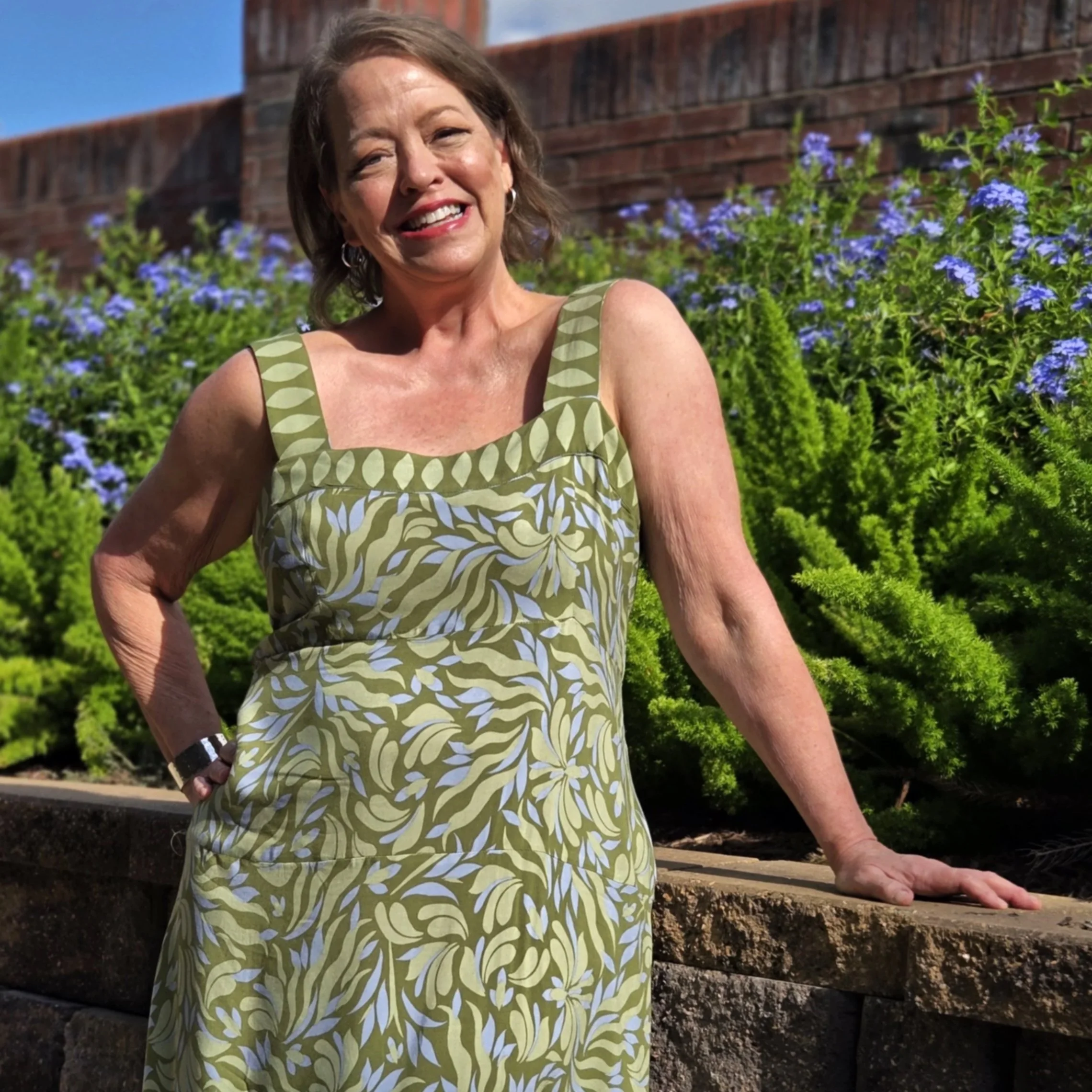 A smiling woman with short brown hair wearing a green and white patterned sleeveless dress, kneeling outdoors on soil in front of a brick wall and purple flowers.