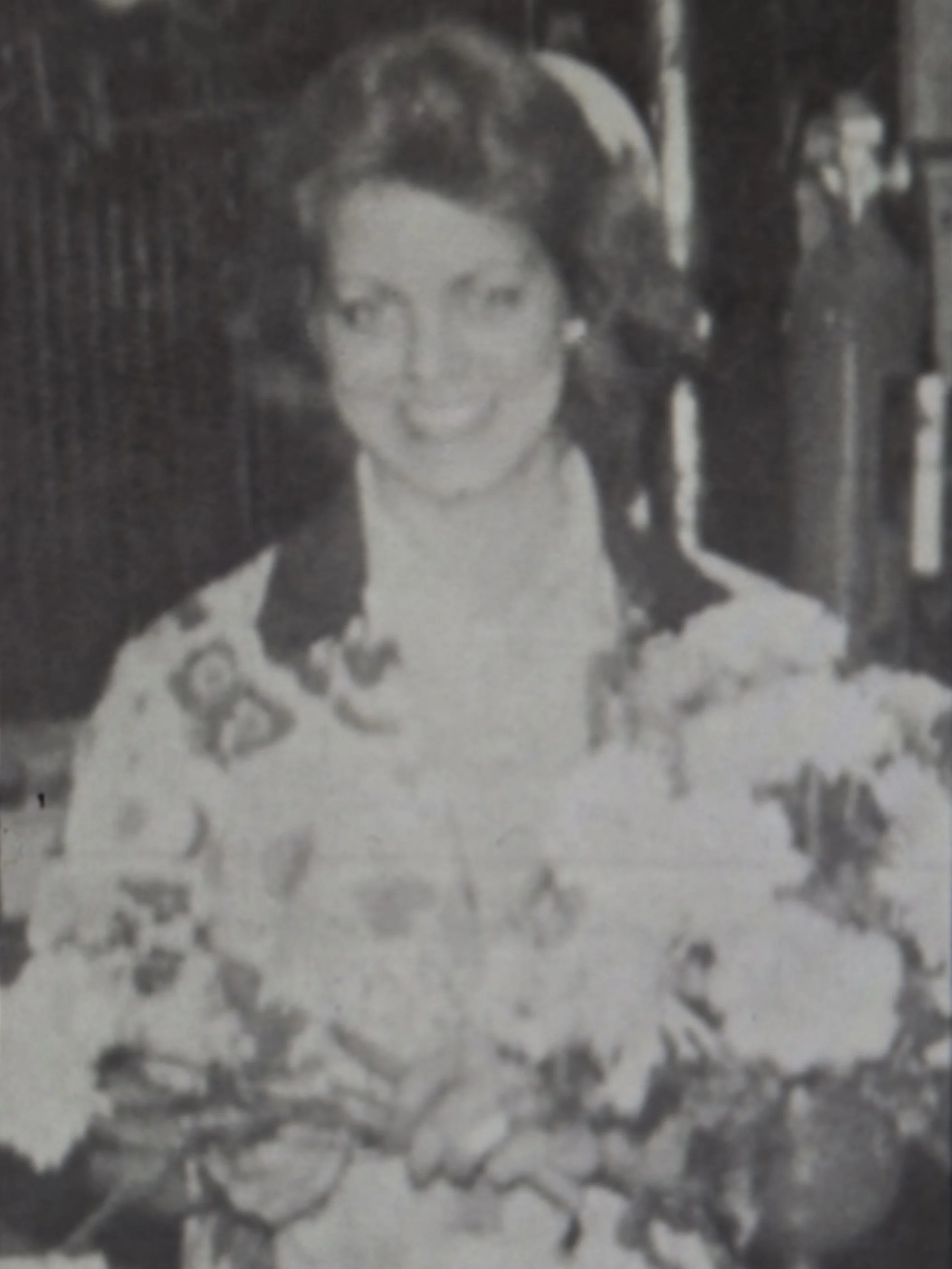 Close-up of a black-and-white photograph of a young girl with dark hair, standing outdoors on a sidewalk, with buildings and a fence in the background.