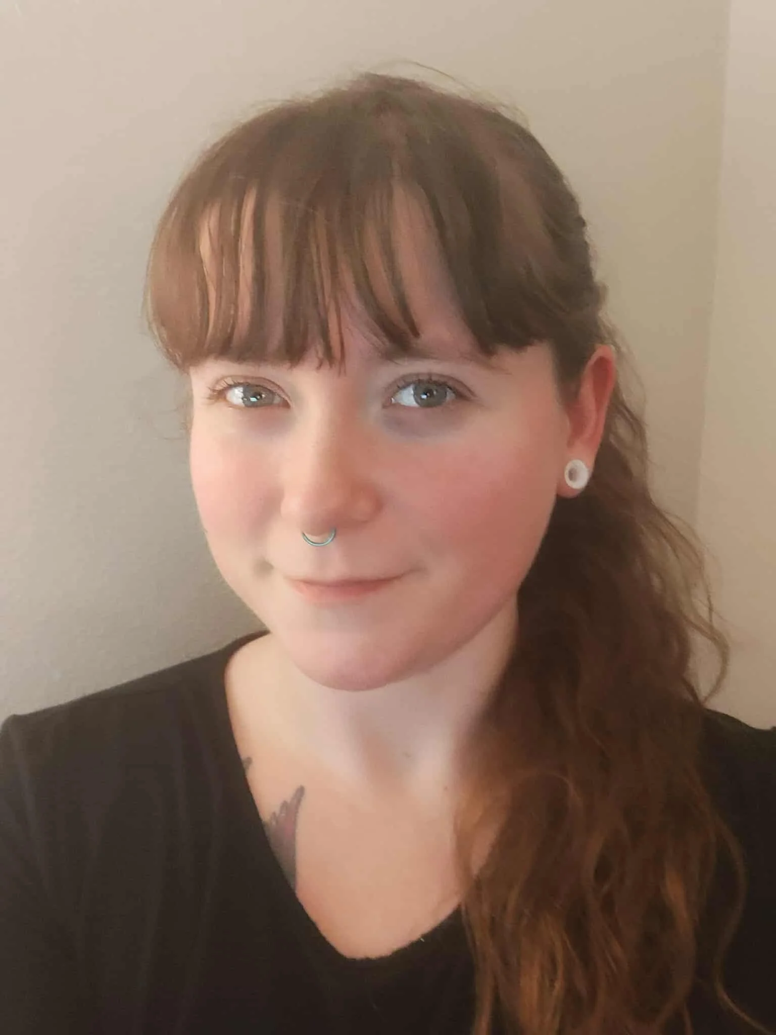 A young woman with long, wavy auburn hair, and blue eyes, wearing a black top, small earrings, and a nose ring, smiling softly at the camera, standing against a plain wall.