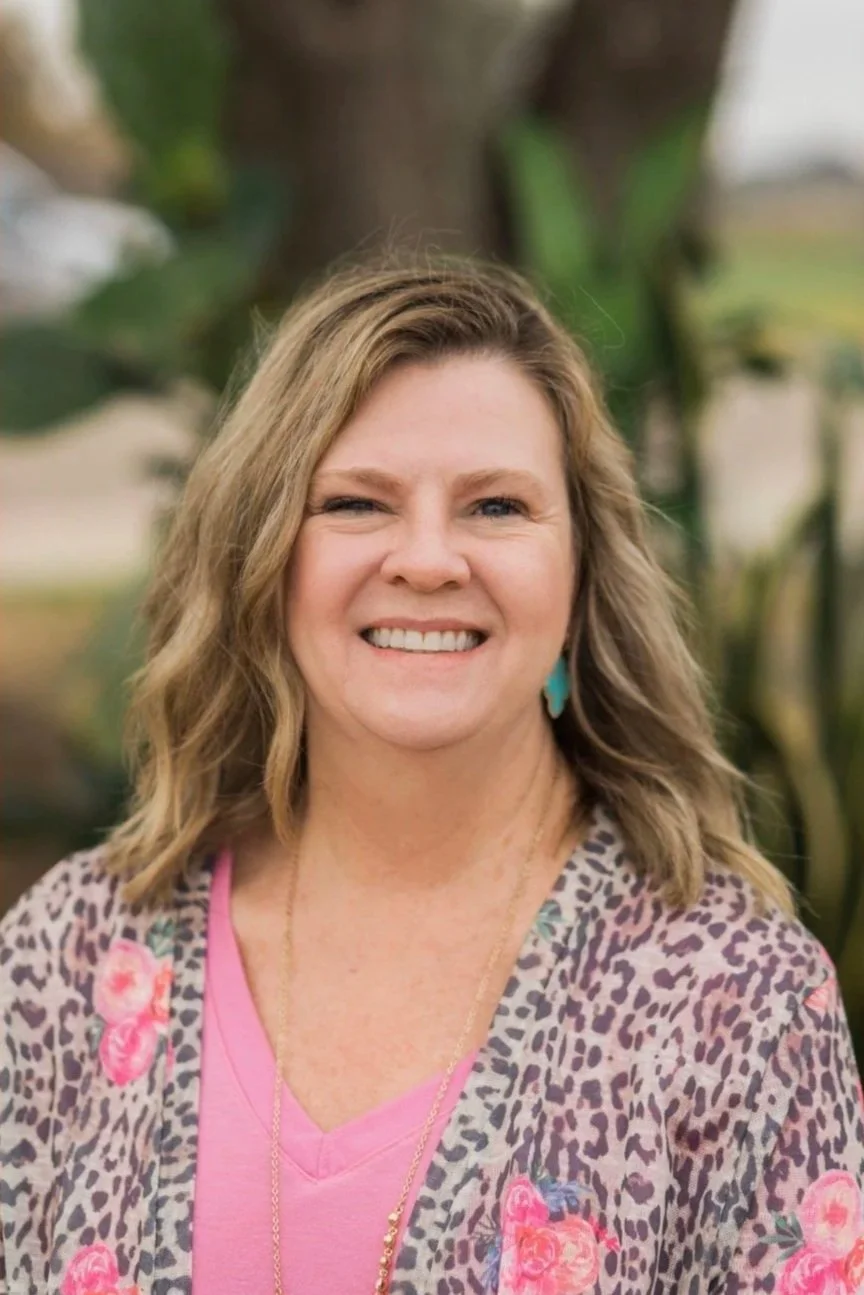 A smiling woman with wavy blonde hair, wearing a leopard print cardigan with pink roses, a pink top, and teal earrings, standing outdoors in front of green foliage.